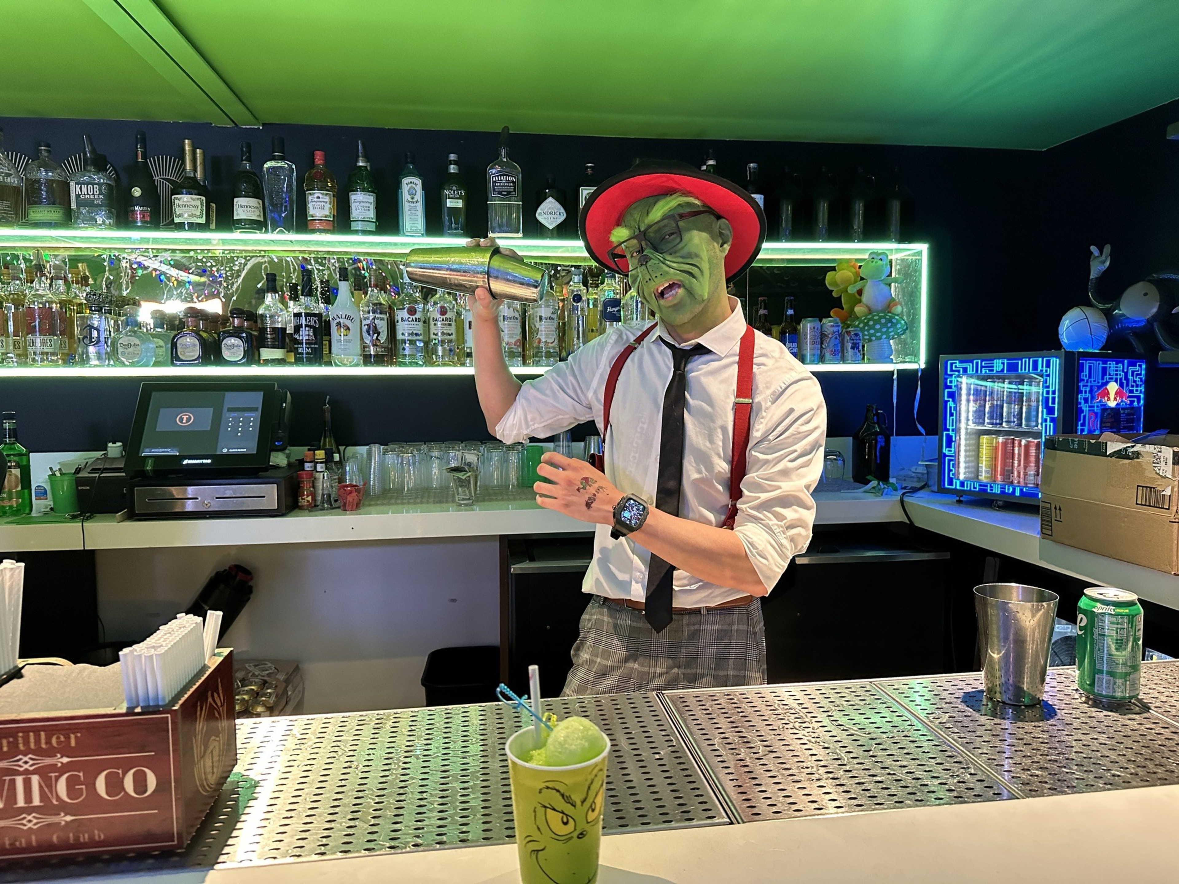 A bartender dressed as Dr. Seuss's the Grinch shakes up a Grinch-themed green cocktail behind a bar.  