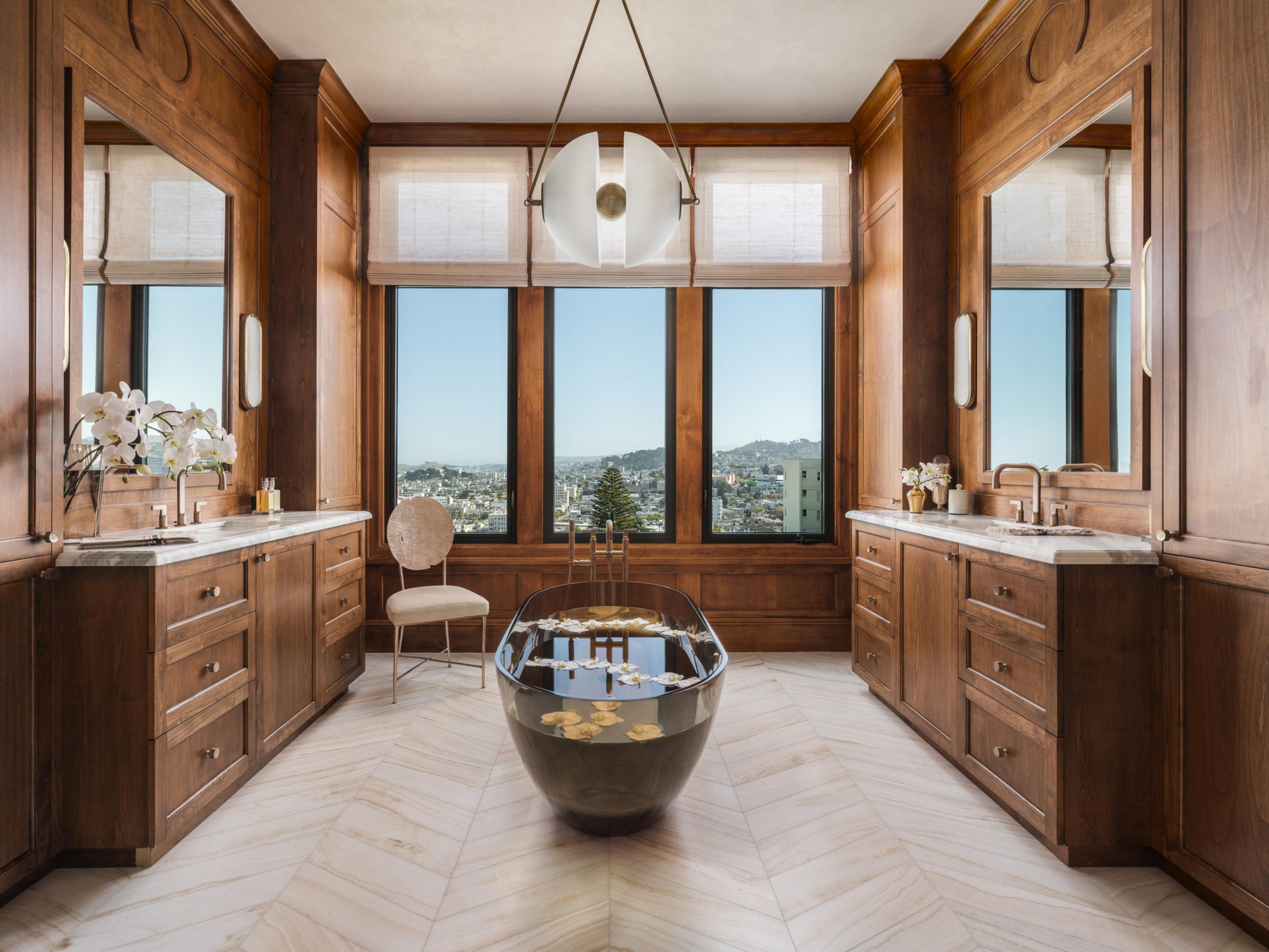 A luxurious bathroom with a black freestanding tub, wood-paneled walls, marble countertops, large mirrors, a beige chair, and a city view through three tall windows.