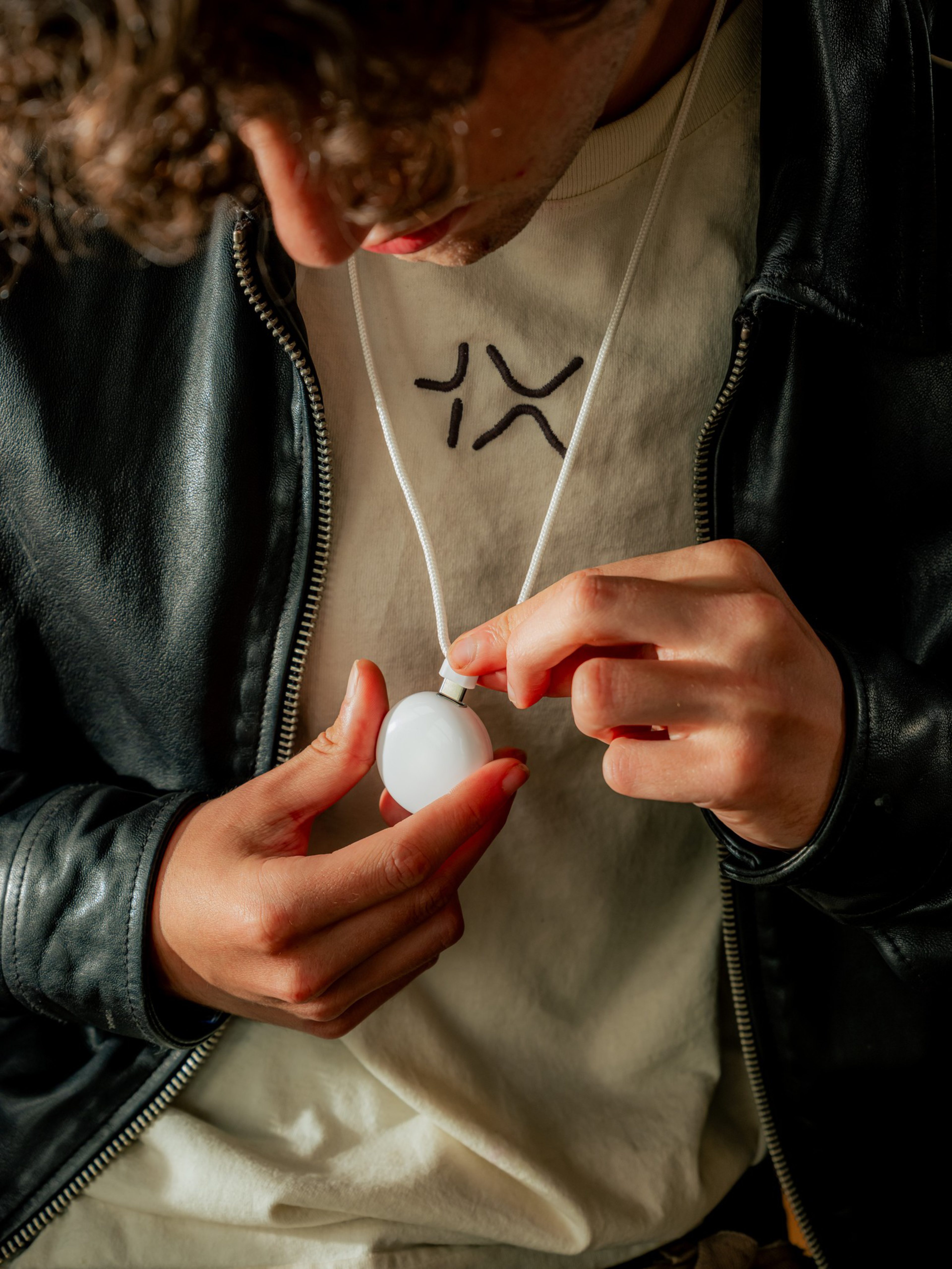 A person wearing a black leather jacket and a cream shirt with a black embroidered design holds a white round object attached to a cord.