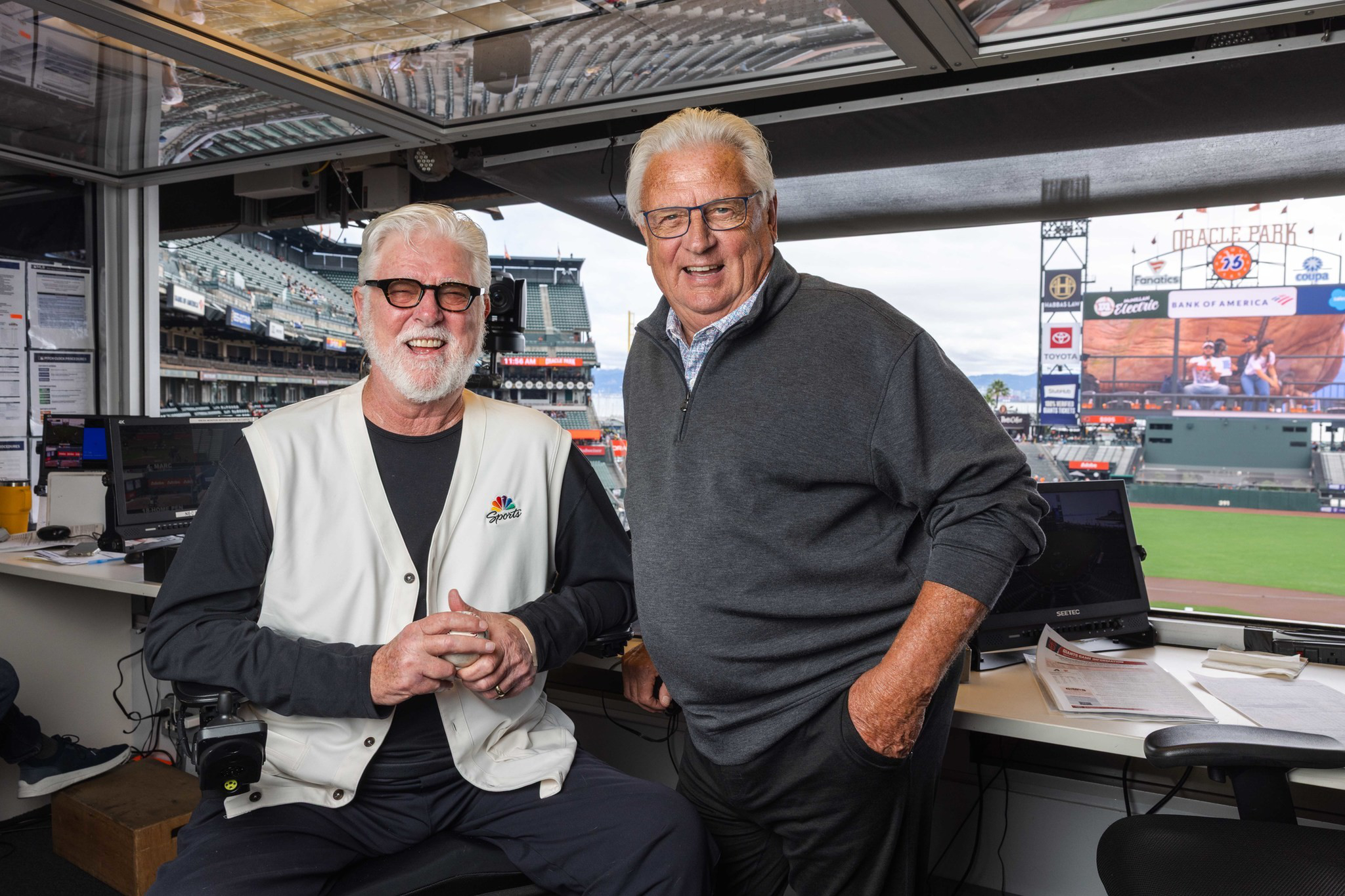 Two older men with gray hair and glasses, one seated wearing an NBC Sports vest, the other standing in a stadium broadcast booth with a baseball field visible.