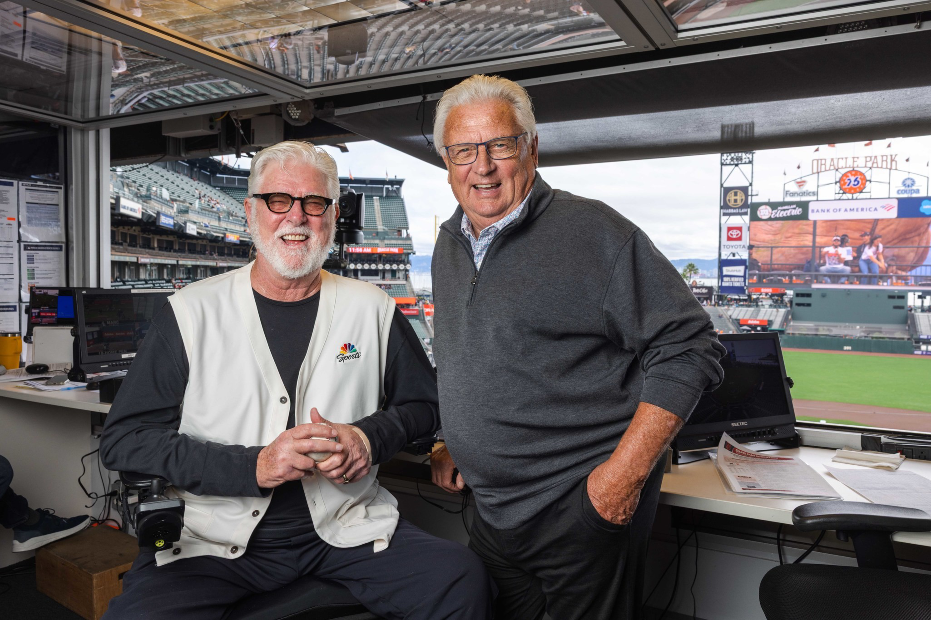 Two older men with gray hair and glasses, one seated wearing an NBC Sports vest, the other standing in a stadium broadcast booth with a baseball field visible.