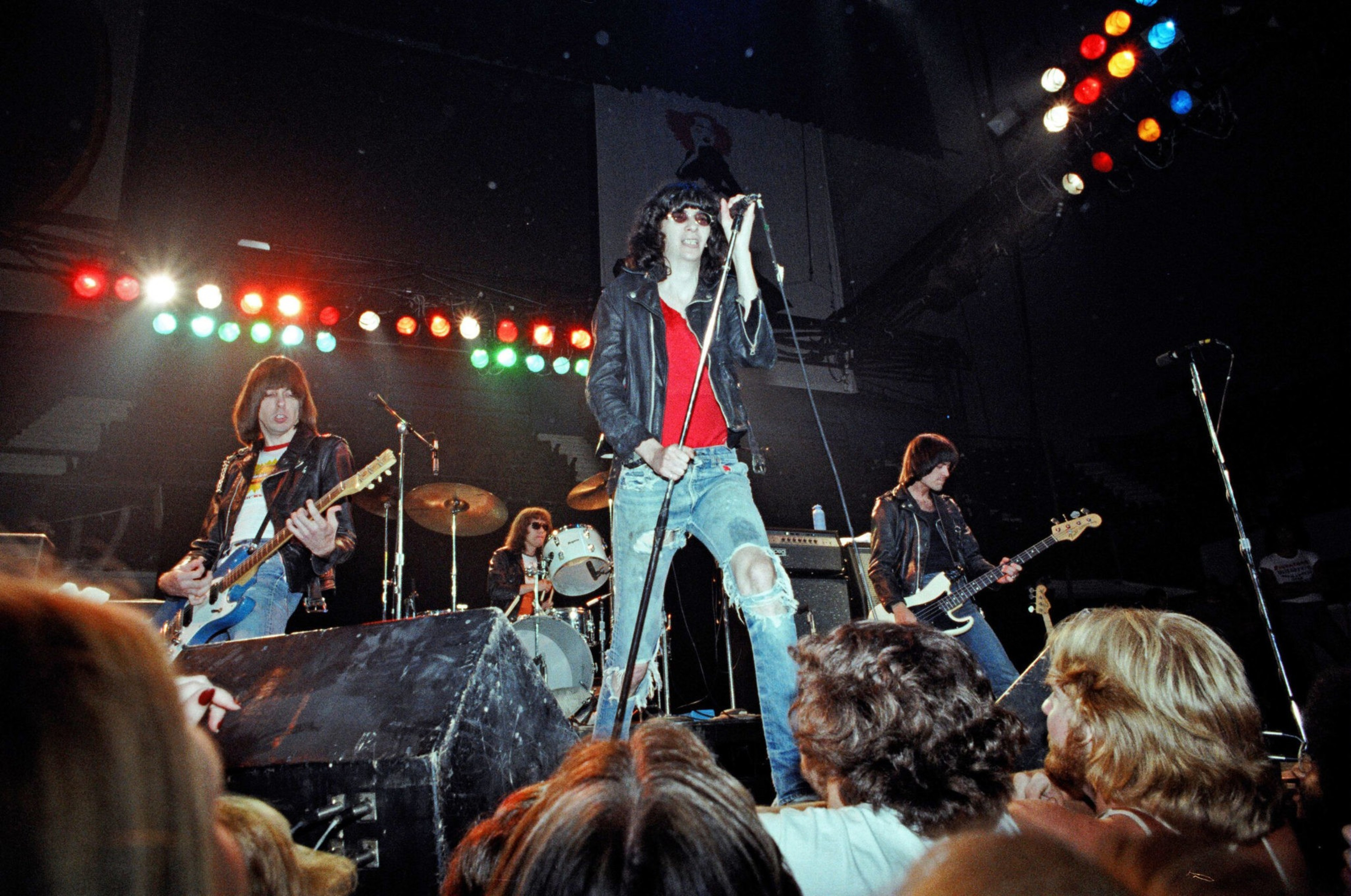 A rock band performs on stage with a singer holding a microphone, flanked by guitarists and a drummer, while a crowd watches closely.