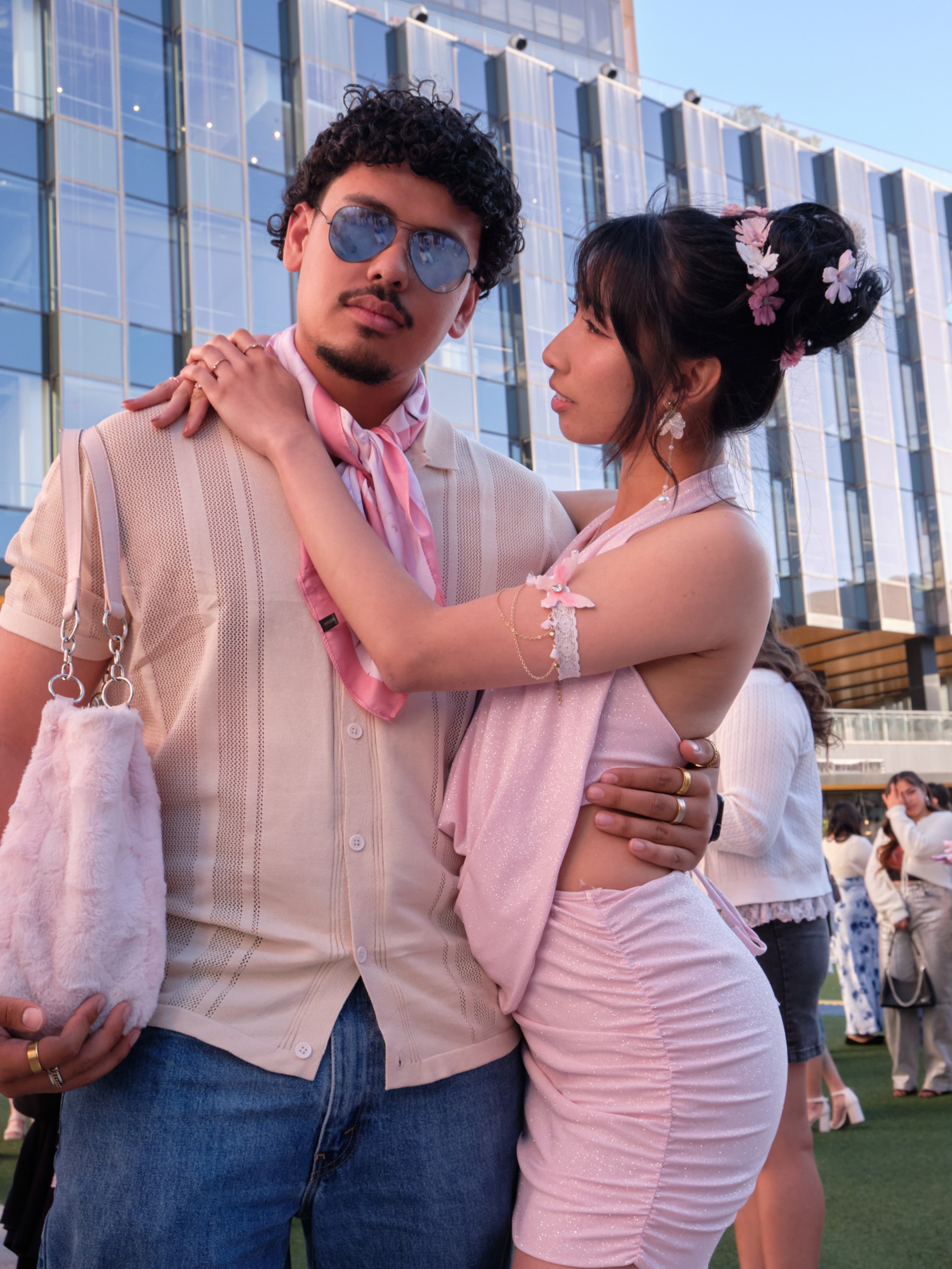 A man wearing sunglasses and a pink scarf is embraced by a woman in a pink dress with floral hair accessories, standing outdoors near a glass building.