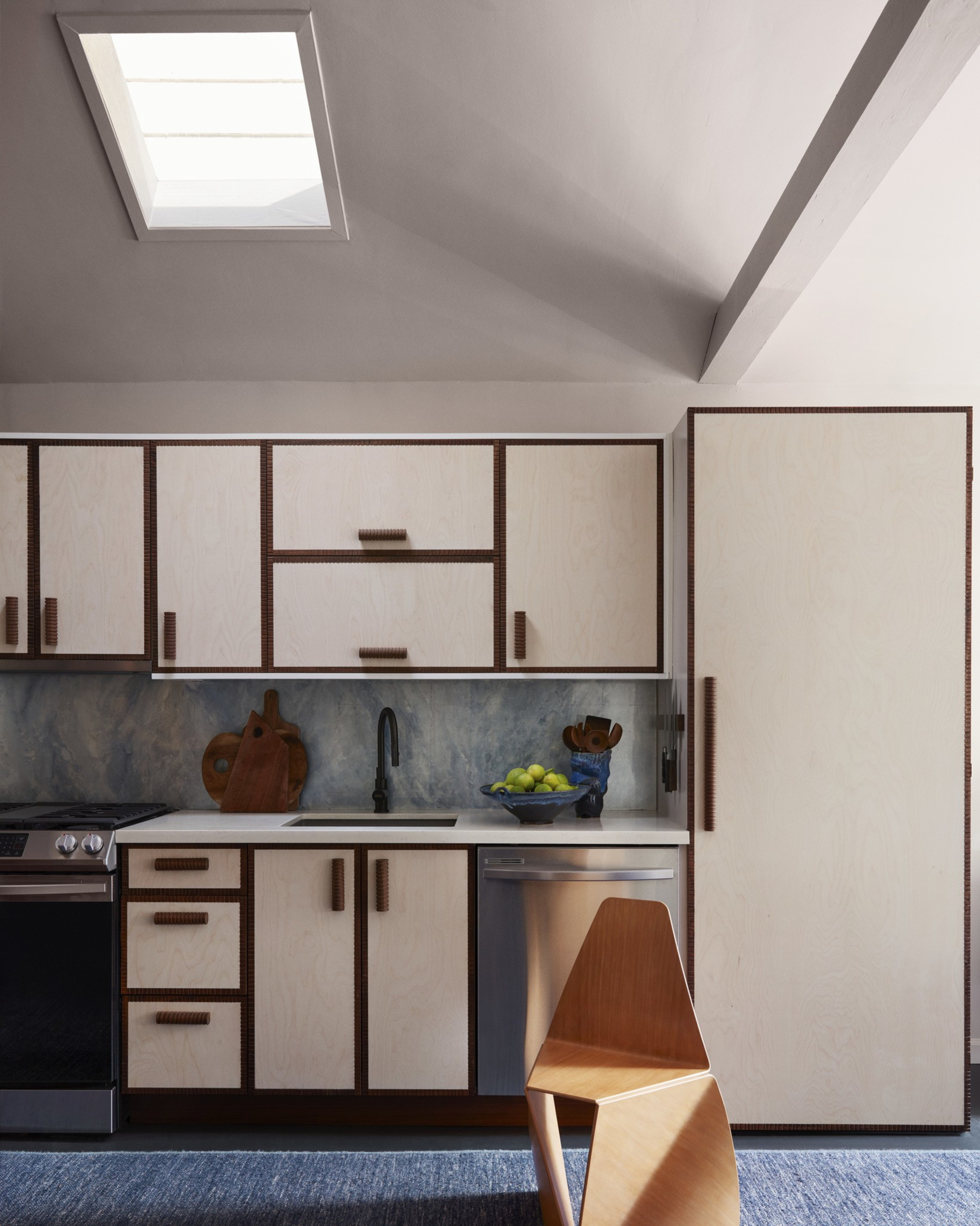 Light wood cabinets with brown trim surround a black faucet and sink under a skylight, next to a stainless dishwasher, with a modern wooden chair on a blue rug.