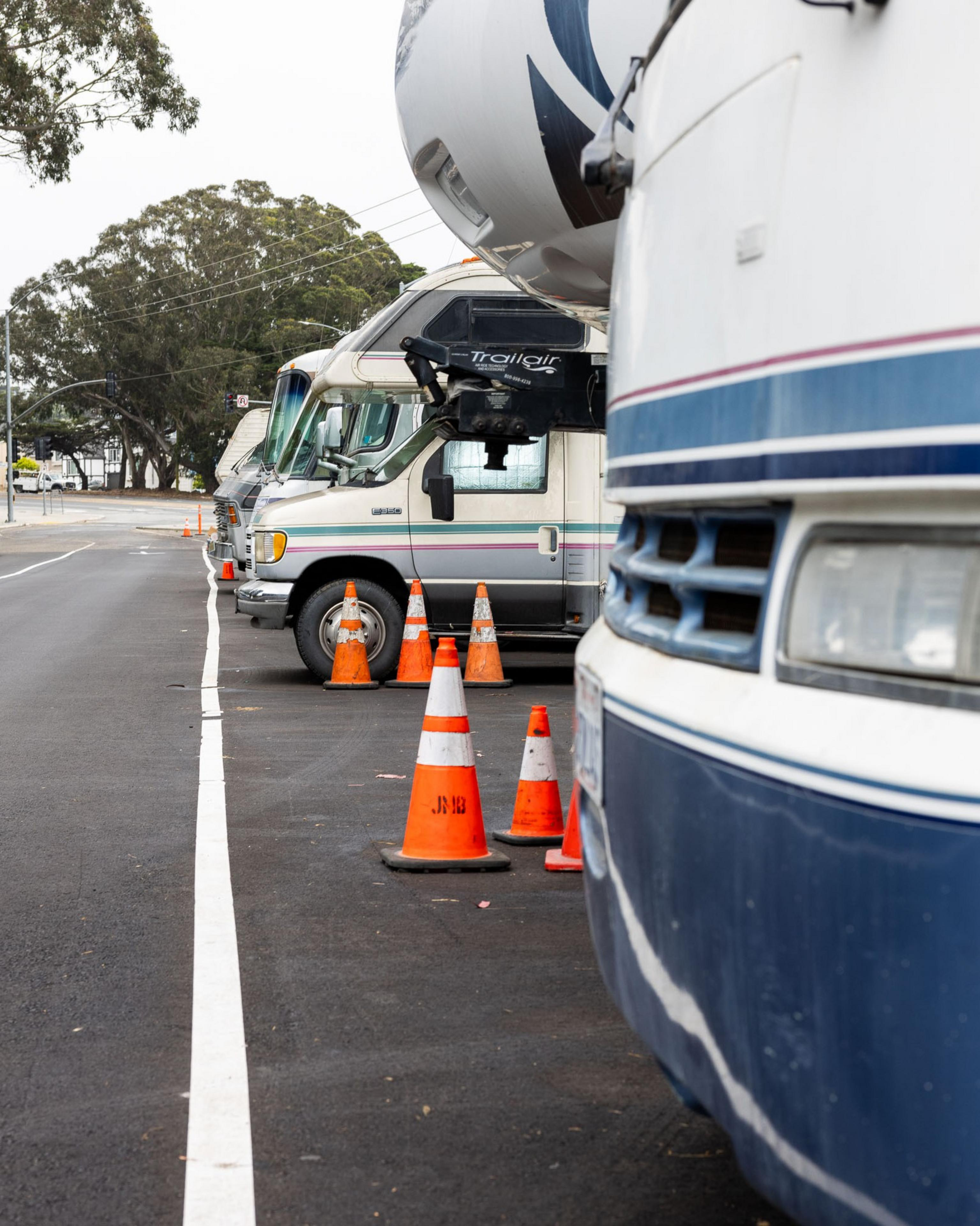 Parked RVs line the side of a street, separated by orange traffic cones. Trees and power lines are visible in the background.