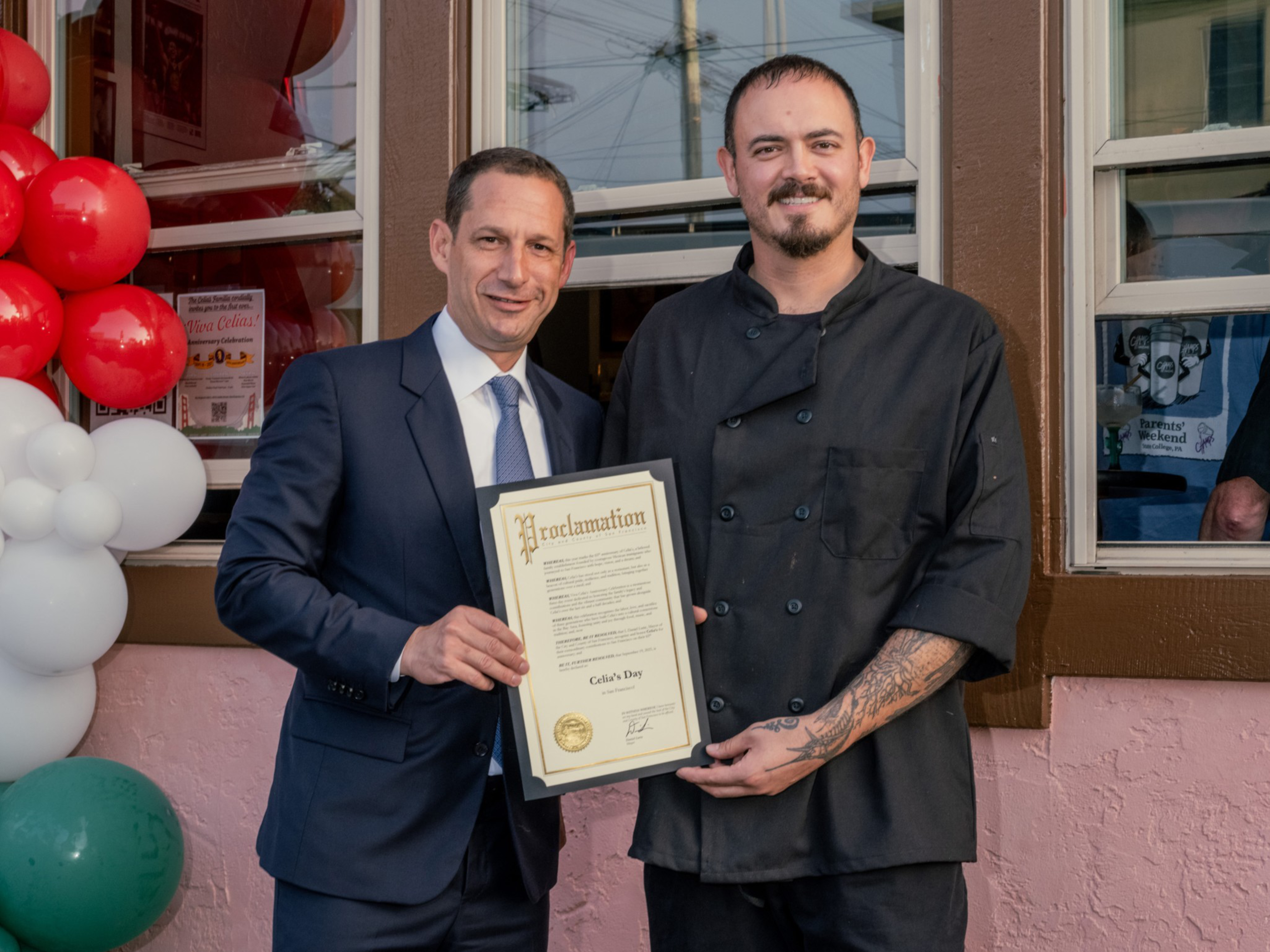 A man in a suit and a chef in black hold a framed proclamation together outside a building decorated with red, white, and green balloons.