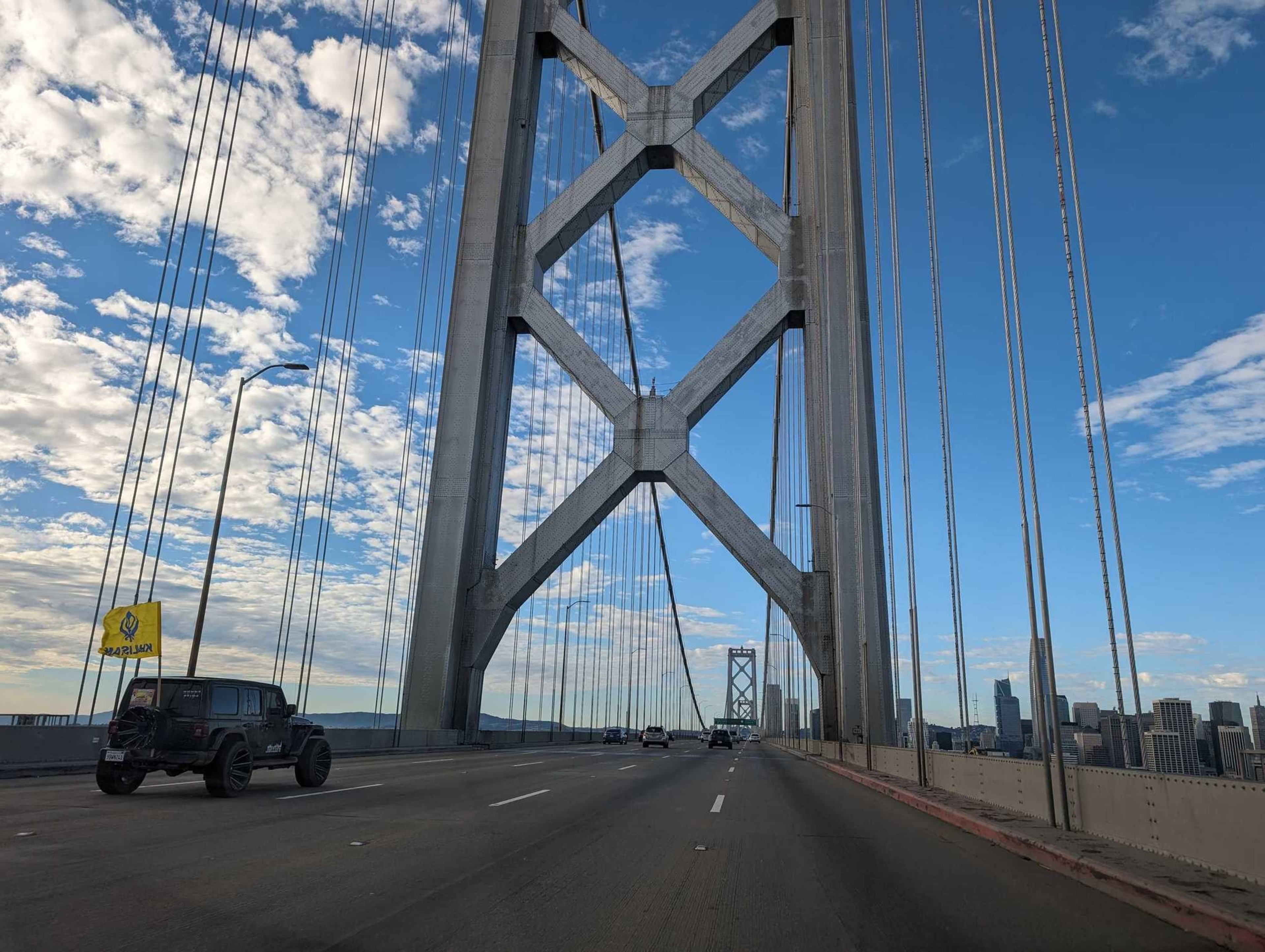 A jeep-style vehicle with a large yellow flag drives along a bridge on its way into a large city
