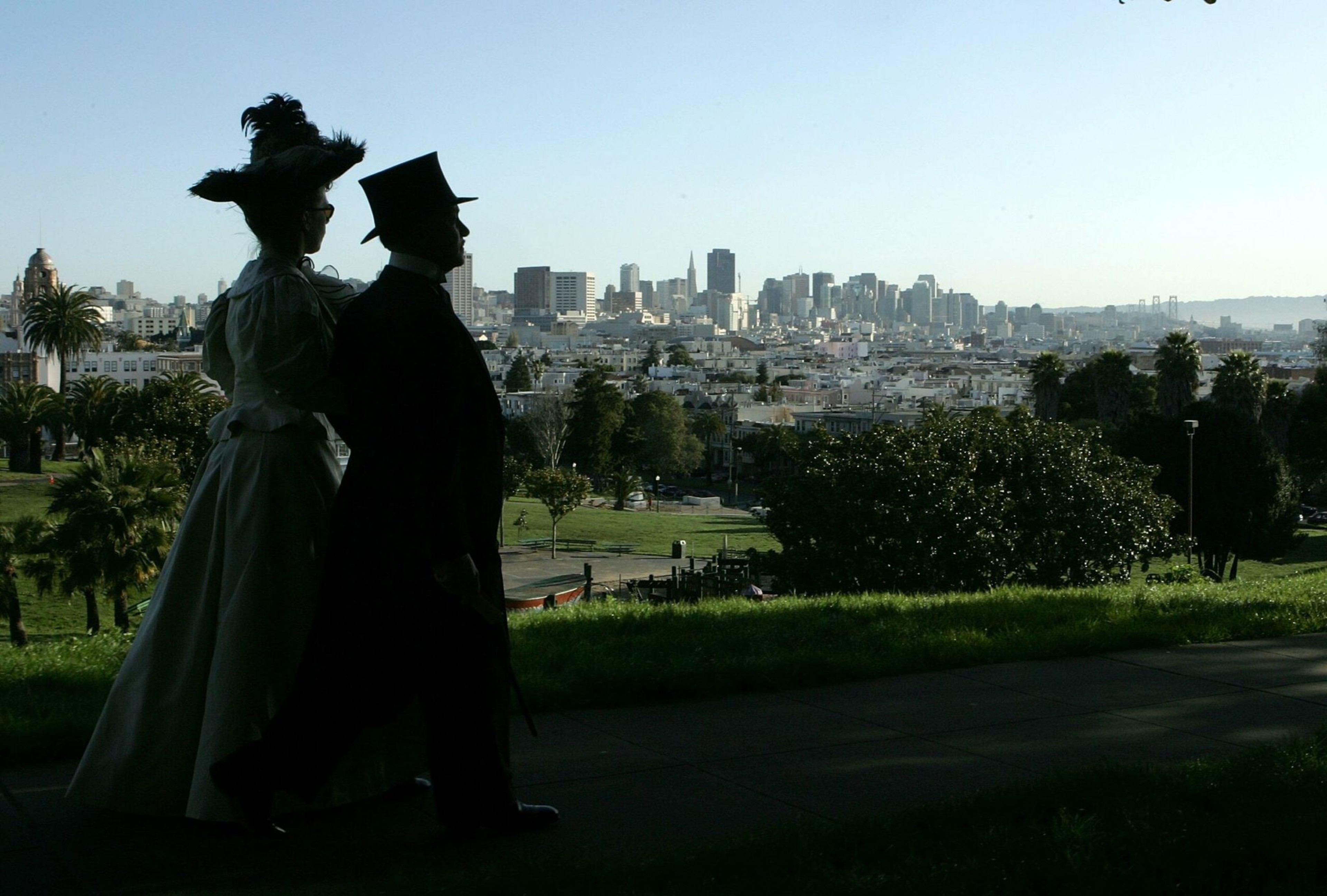 A couple dressed in Victorian-era clothing walks along a park path with a city skyline and clear sky in the background.