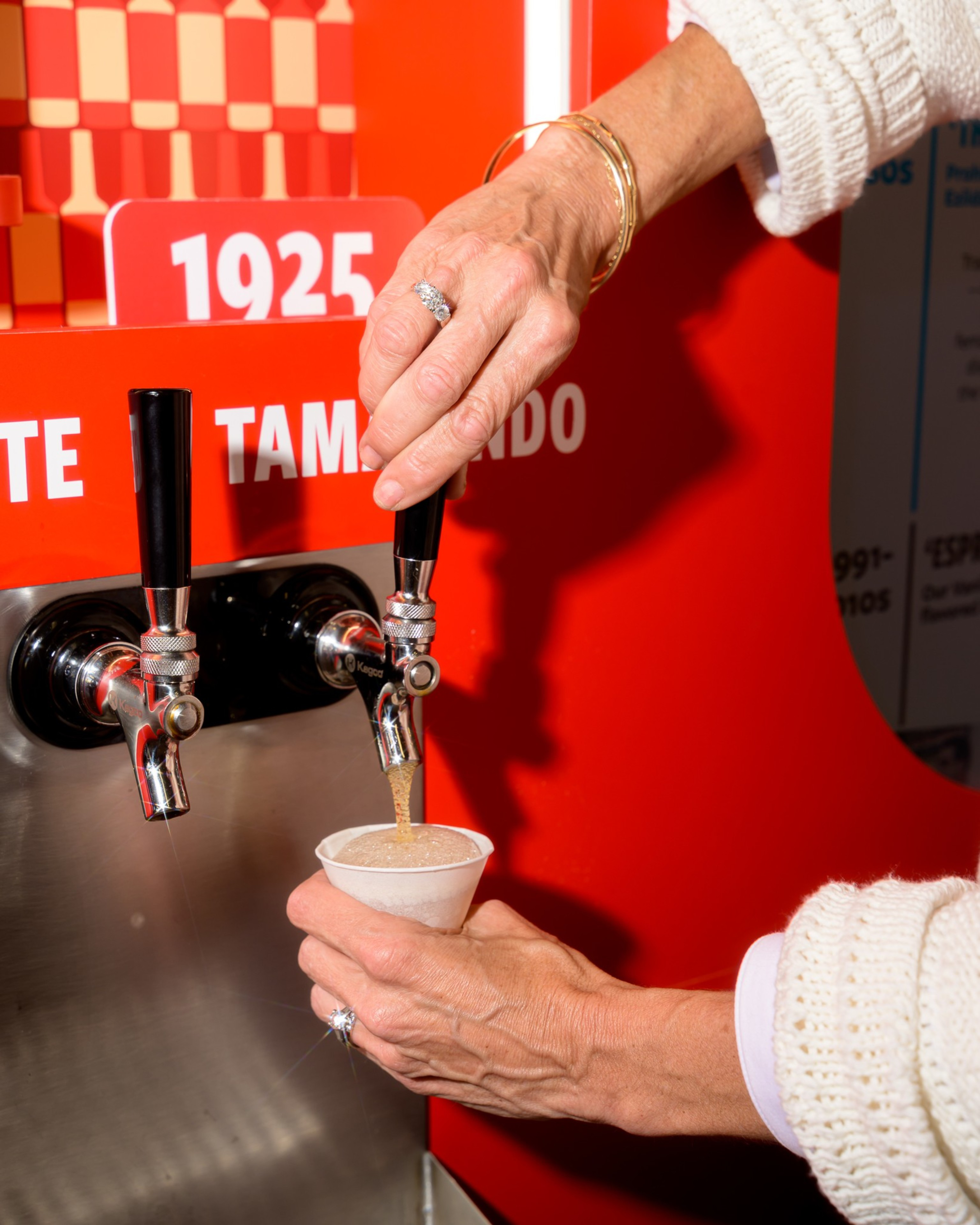 A person pours tamarindo-flavored drink from a dispenser into a white paper cup. Their hand is adorned with a ring, and the dispenser is shiny and red.