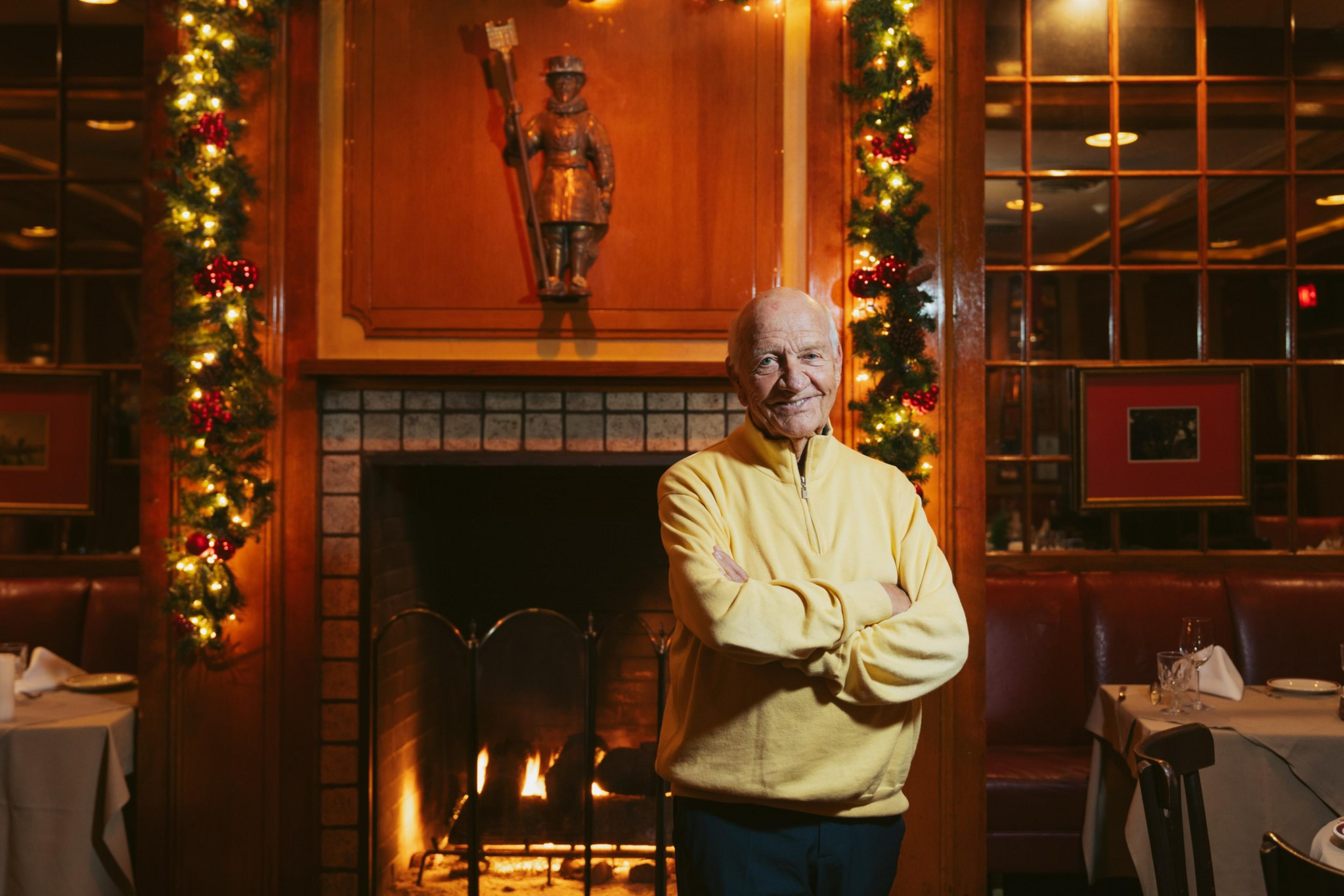 An elderly man in a yellow sweater stands smiling with arms crossed by a lit fireplace decorated with Christmas garlands in a cozy restaurant.