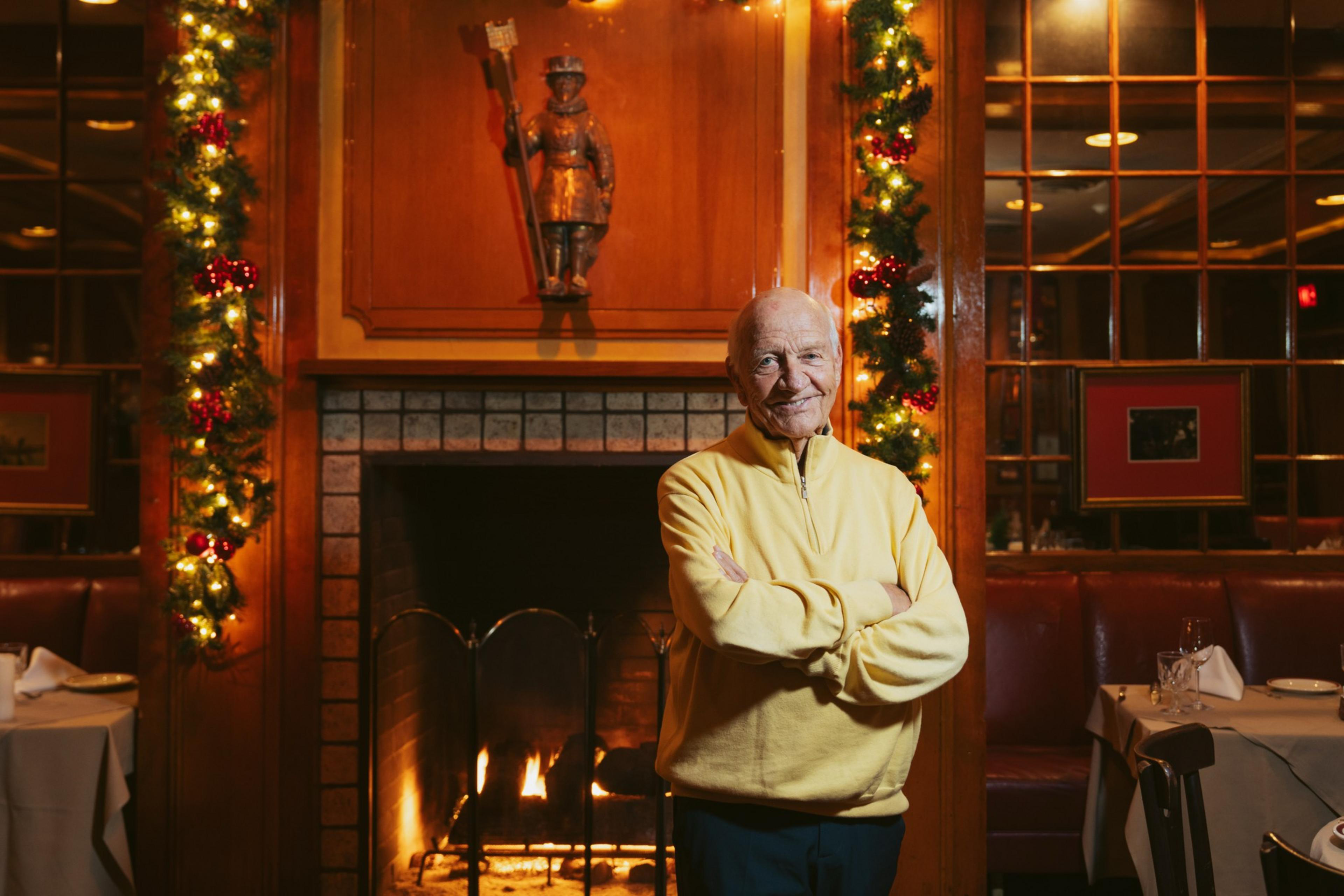 An elderly man in a yellow sweater stands smiling with arms crossed by a lit fireplace decorated with Christmas garlands in a cozy restaurant.
