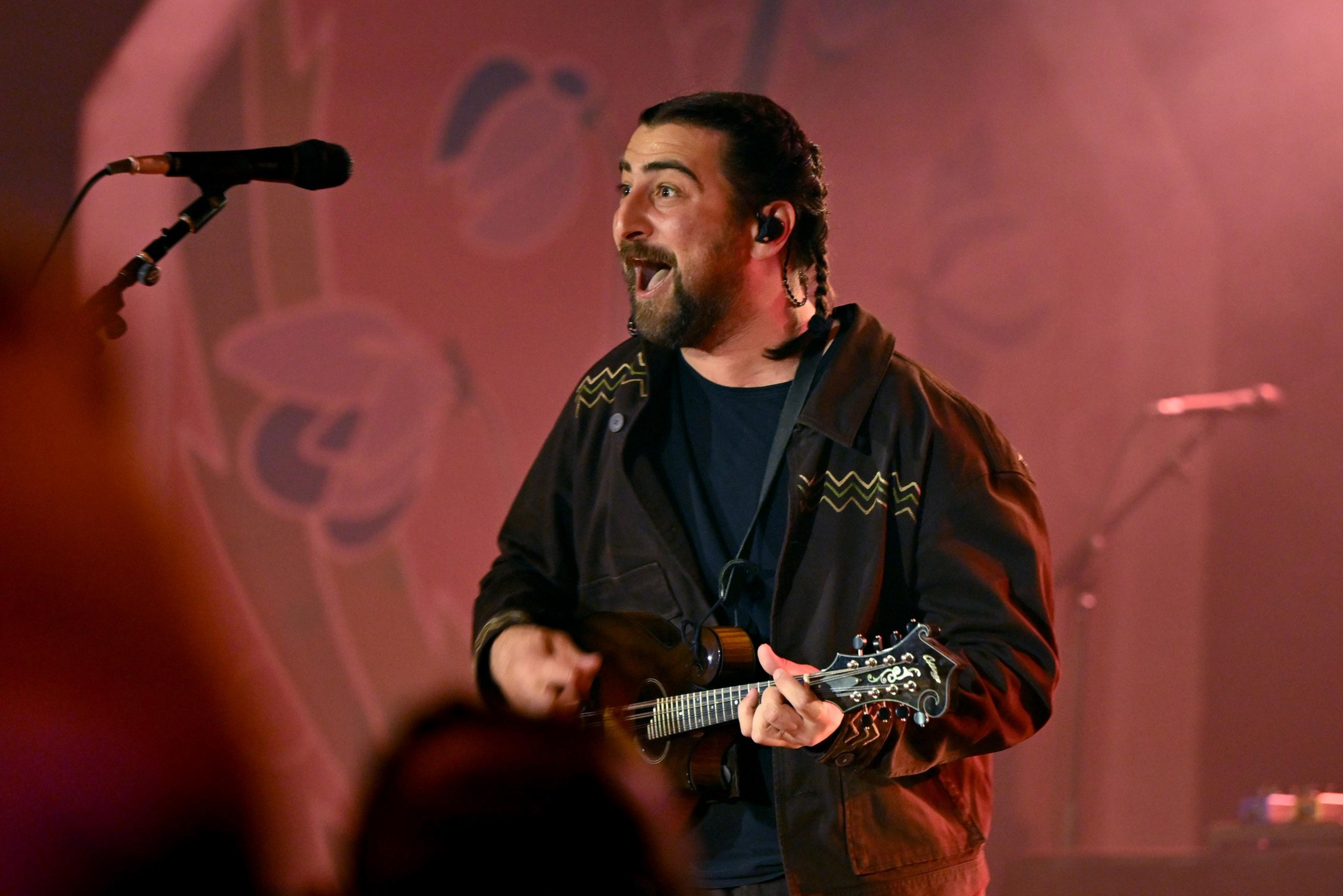 A musician enthusiastically plays a string instrument on stage, with a microphone nearby. The backdrop is warmly lit with abstract patterns.