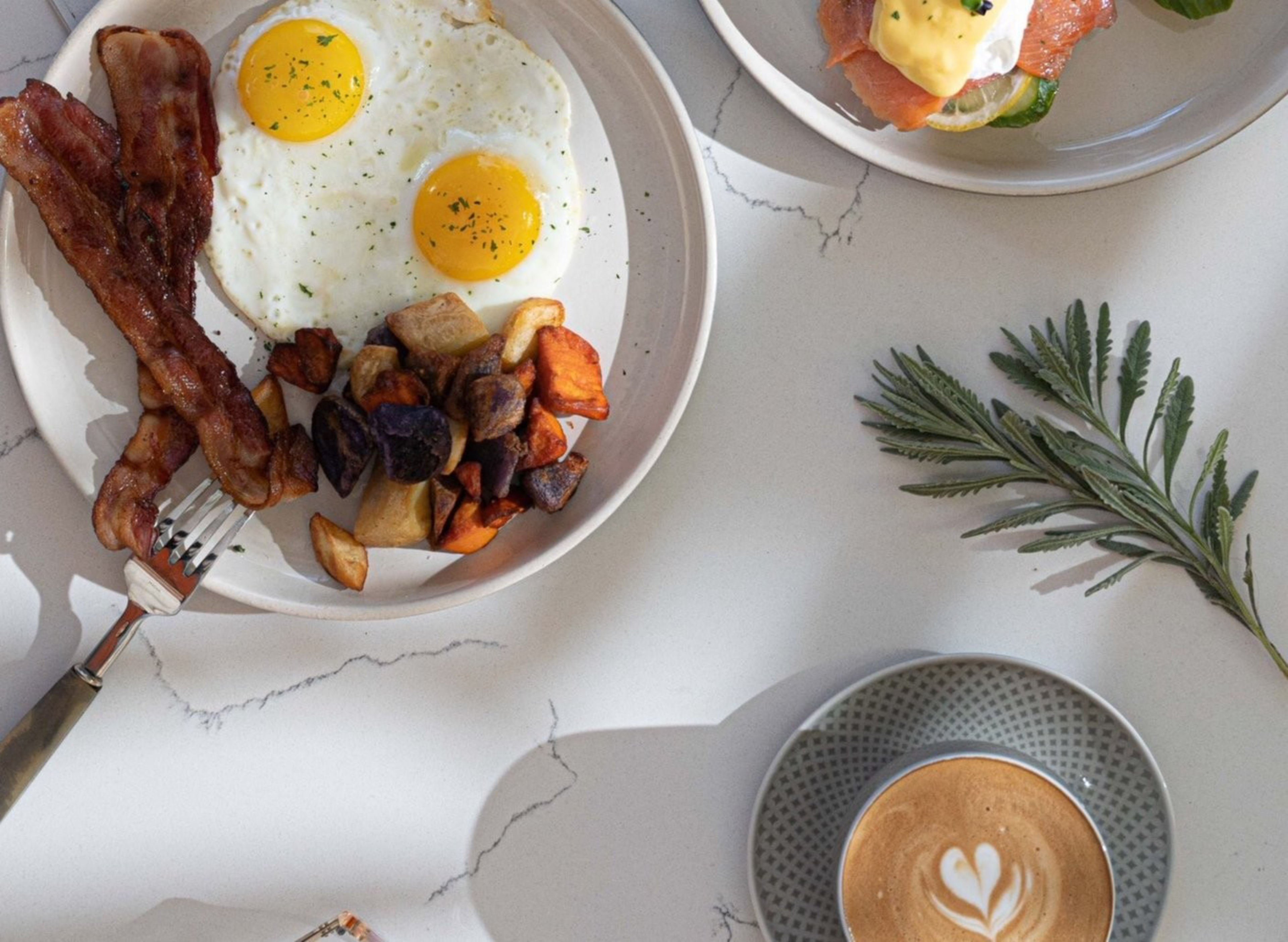 Two sunny-side-up eggs, two strips of bacon, roasted potatoes, smoked salmon with hollandaise sauce on toast, a cup of coffee with latte art, and a sprig of herbs.