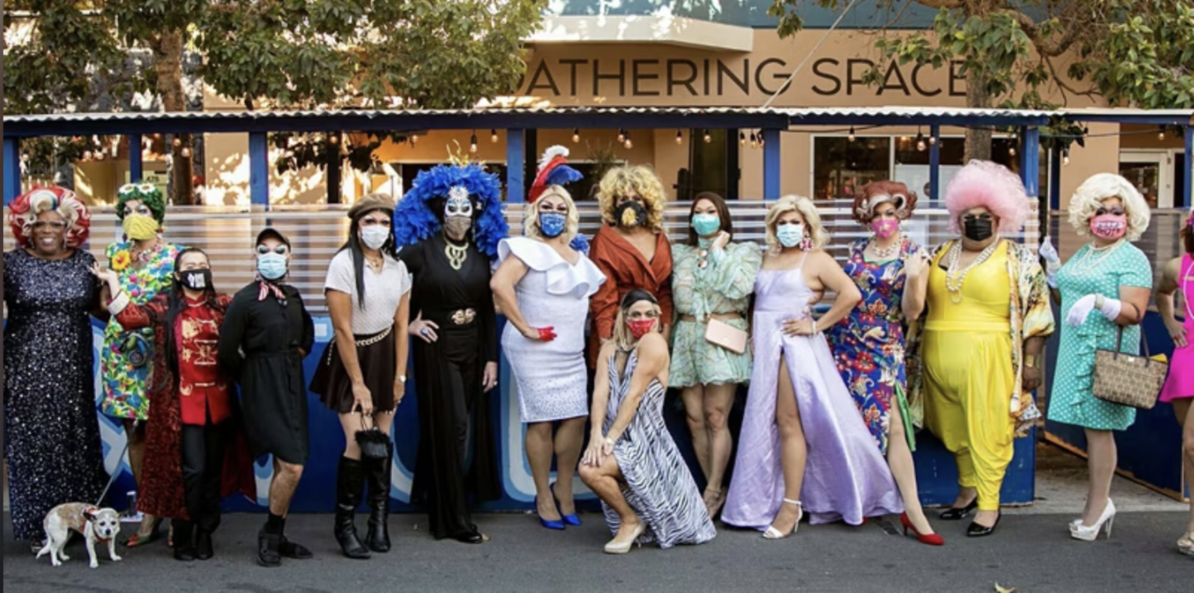 A diverse group in colorful, elaborate costumes and face masks stands outdoors in front of a building labeled "Gathering Space," posing for a photo.