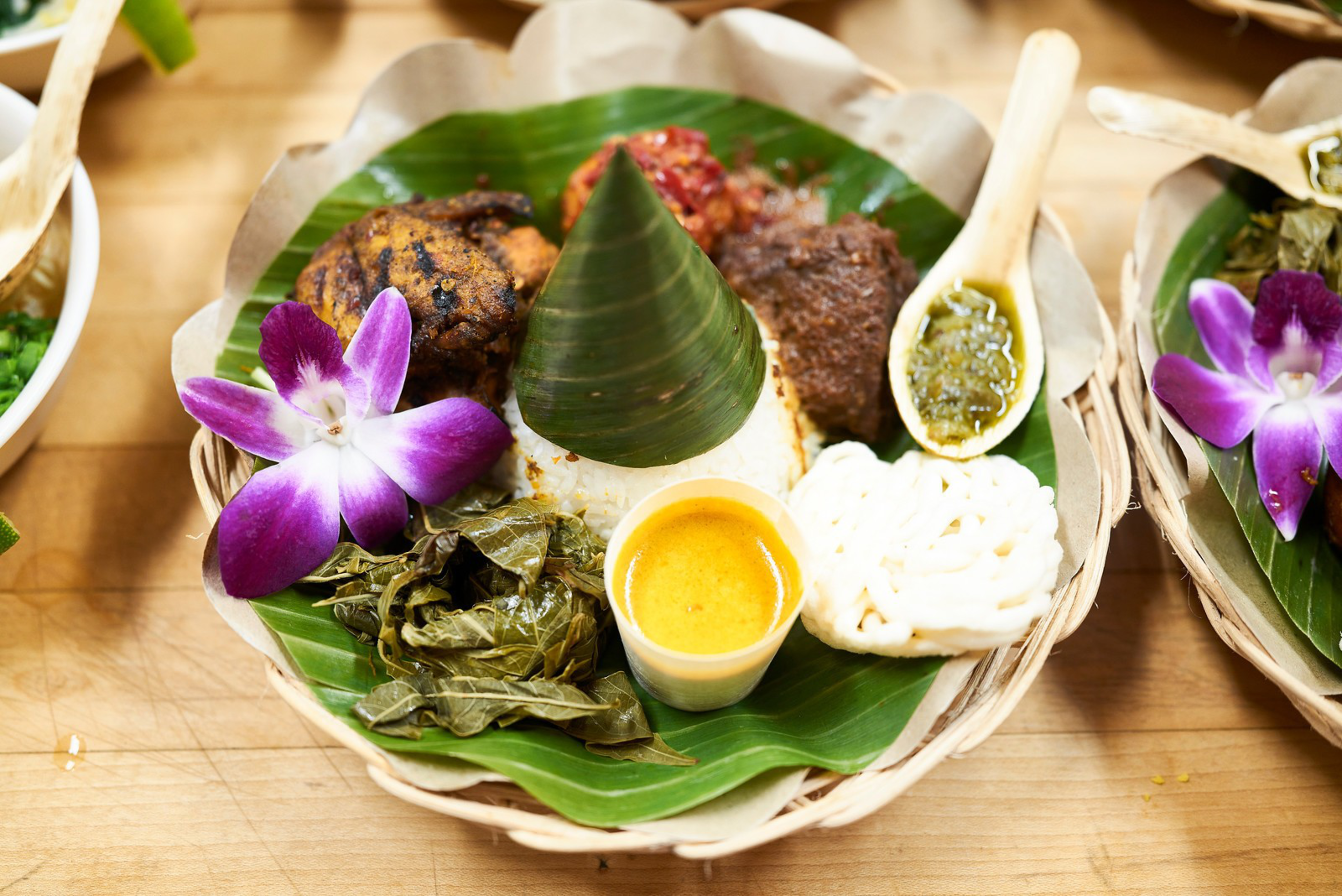 A woven basket contains rice topped with a banana leaf cone, grilled meat, wrapped greens, a small cup of yellow sauce, a white cracker, a spoon of green sauce, and a purple orchid.