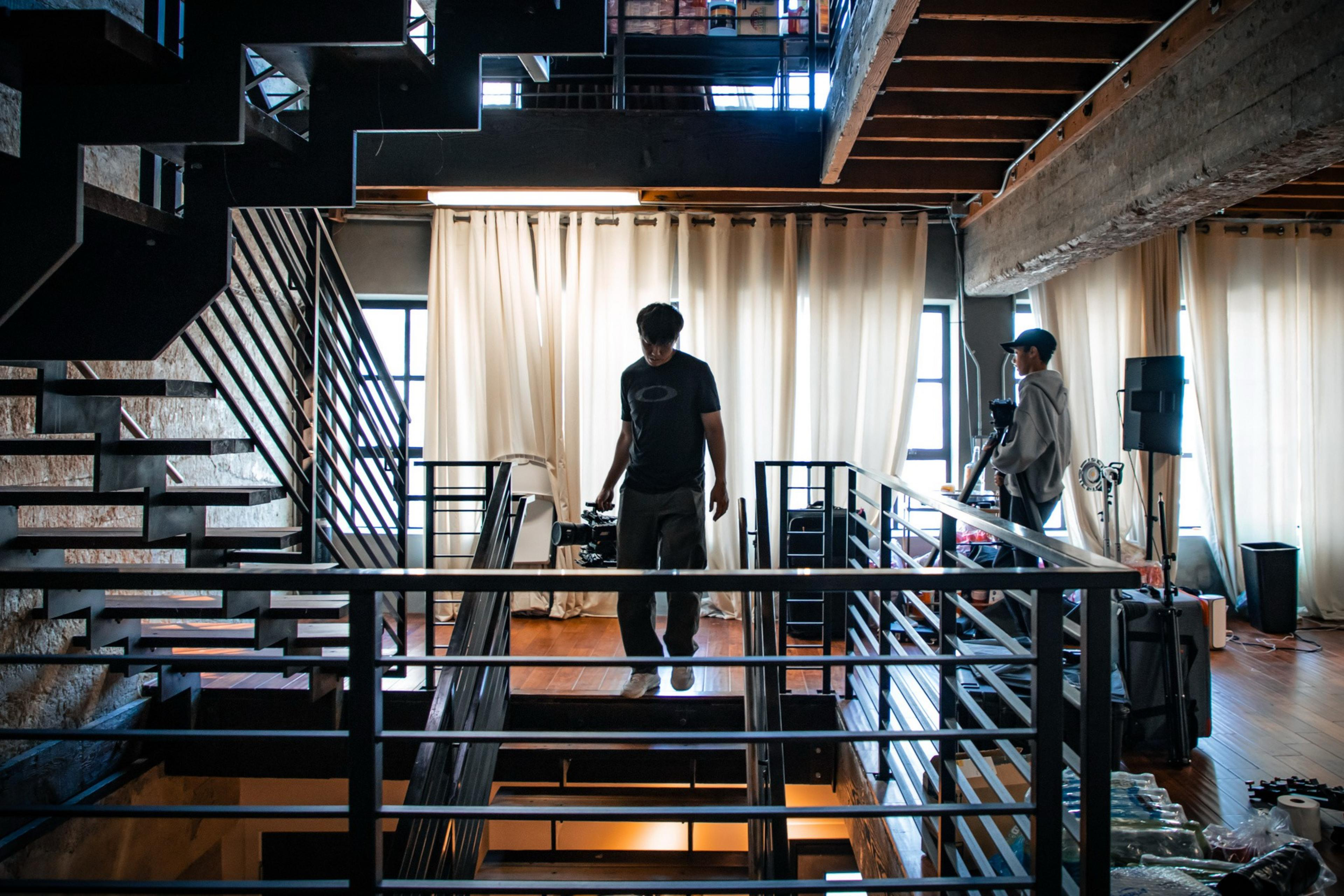 Two people stand in a modern loft with wooden floors and metal railings. One holds a camera, the other stands by equipment near large windows with curtains.