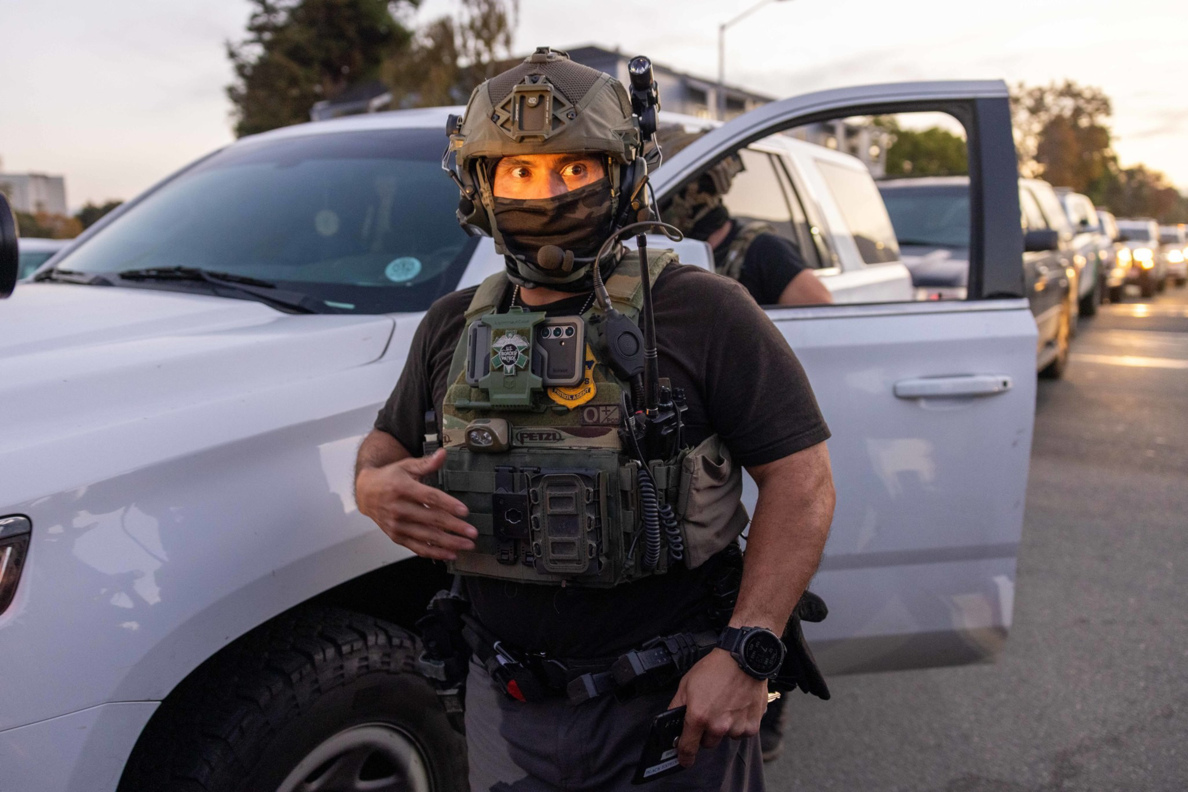 A person wearing tactical gear, a helmet, and a mask stands beside a white vehicle with its door open during dusk or dawn.