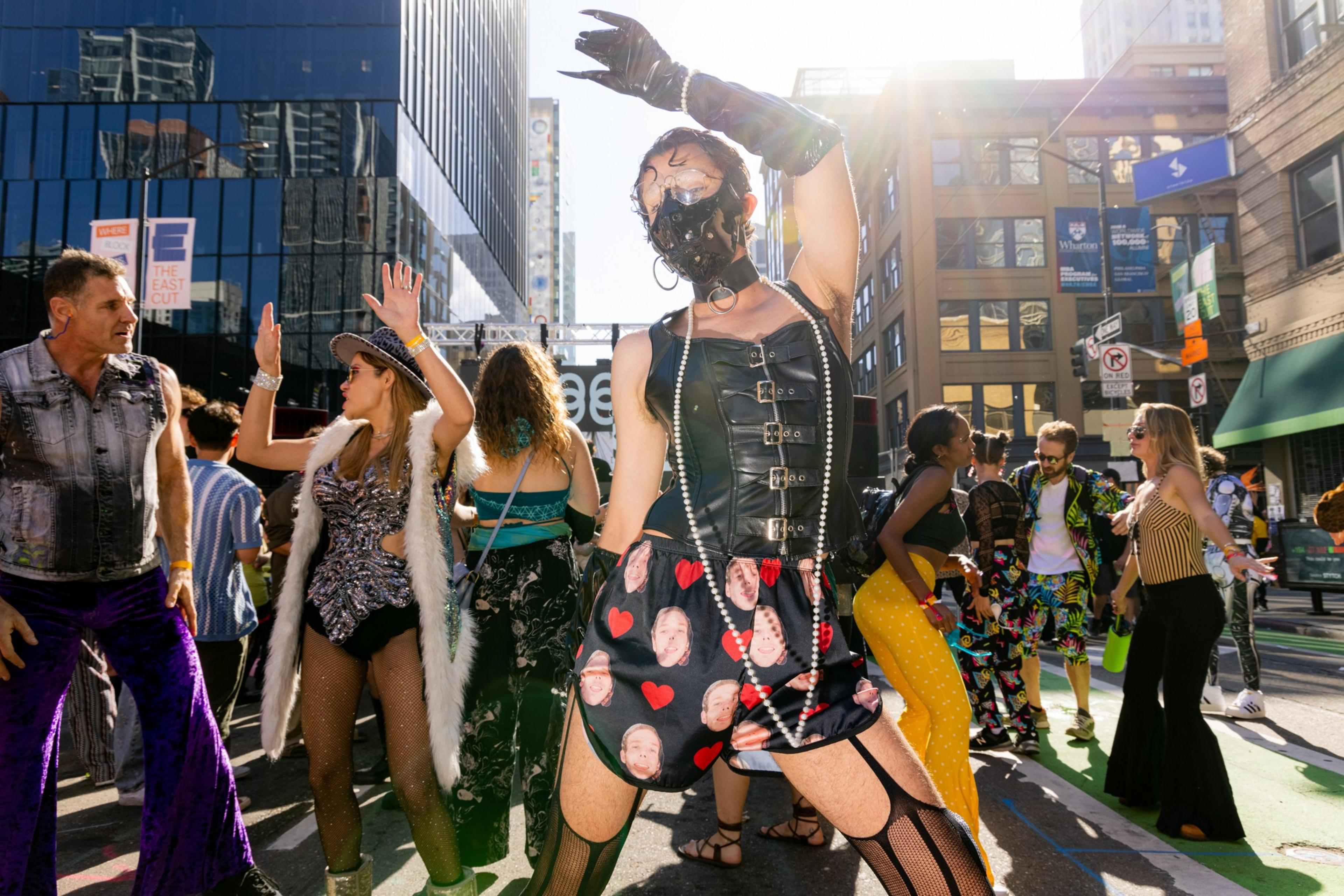 A lively street scene shows people in colorful, eclectic costumes dancing and celebrating in a city, with tall buildings in the background under bright sunlight.