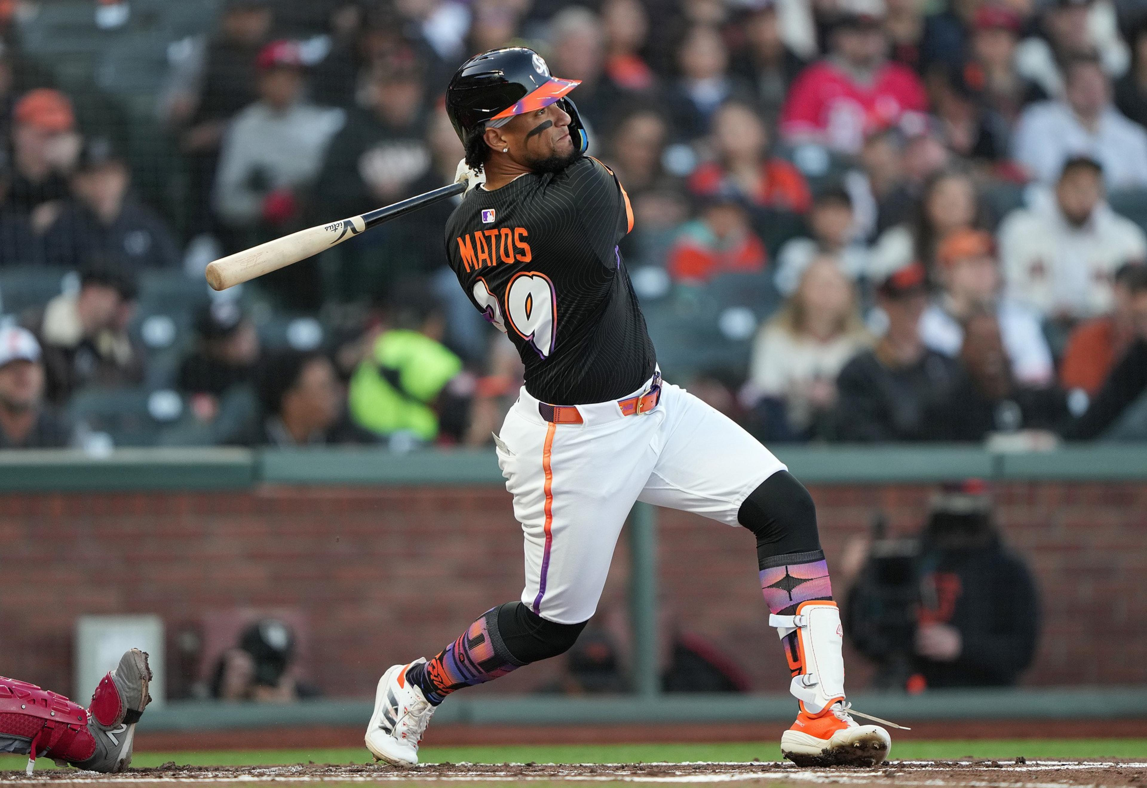 A baseball player in a black and orange uniform is swinging a bat. The player's name, "Matos," and the number 9 are visible on the jersey. The audience is blurred in the background.