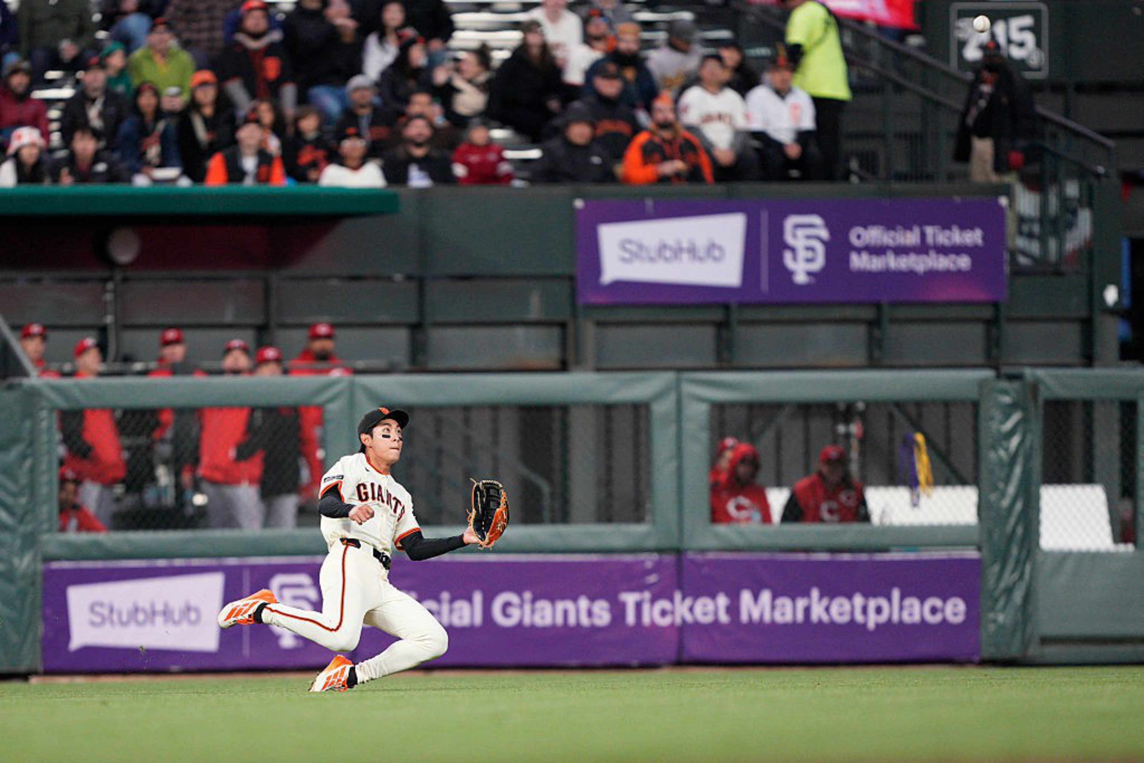 A baseball player in a Giants uniform is mid-action with a glove extended to catch a ball on a field. Spectators and team members are visible in the background.