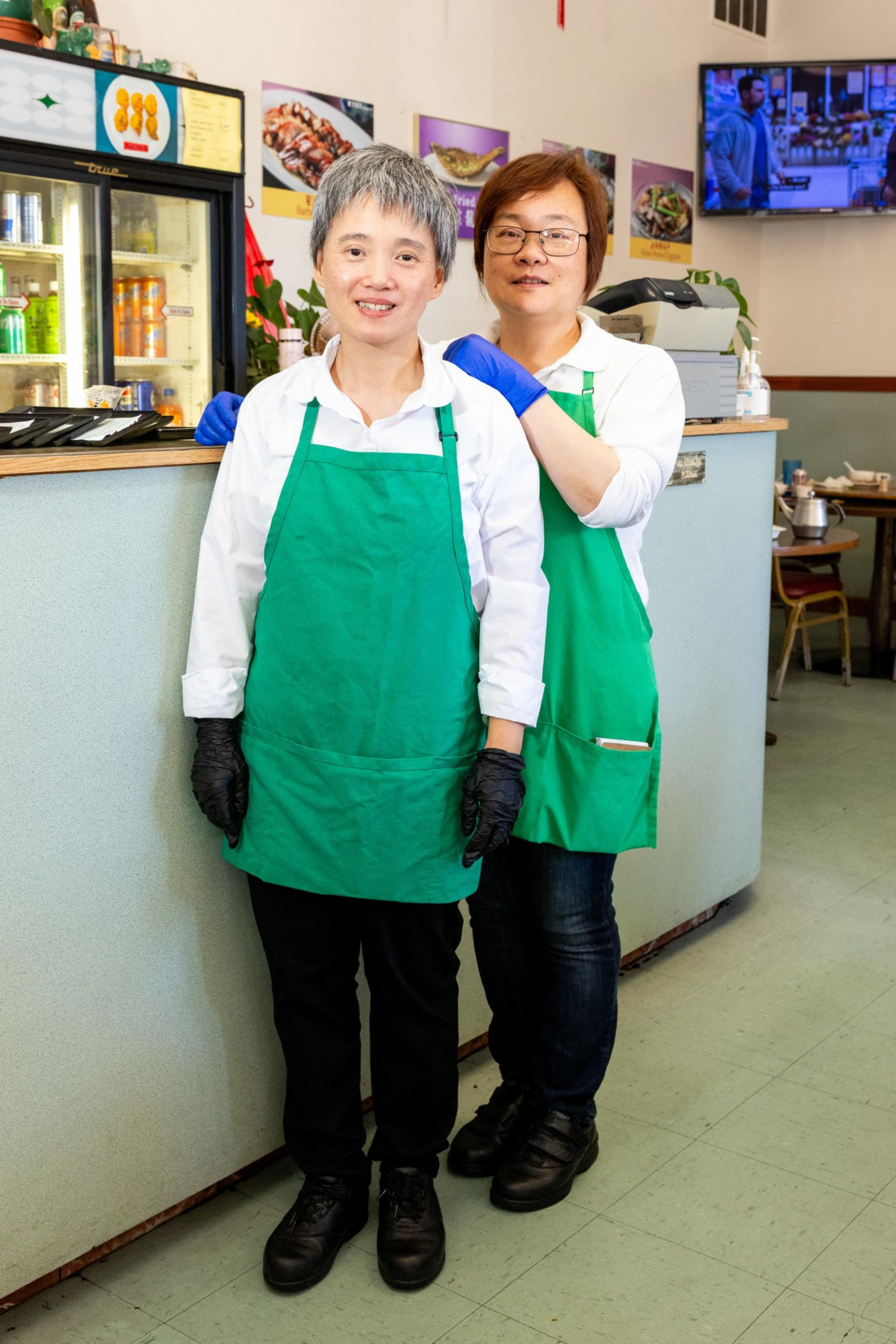 Two women wearing green aprons and gloves stand side by side inside a restaurant, smiling near a counter with drinks and food pictures behind them.