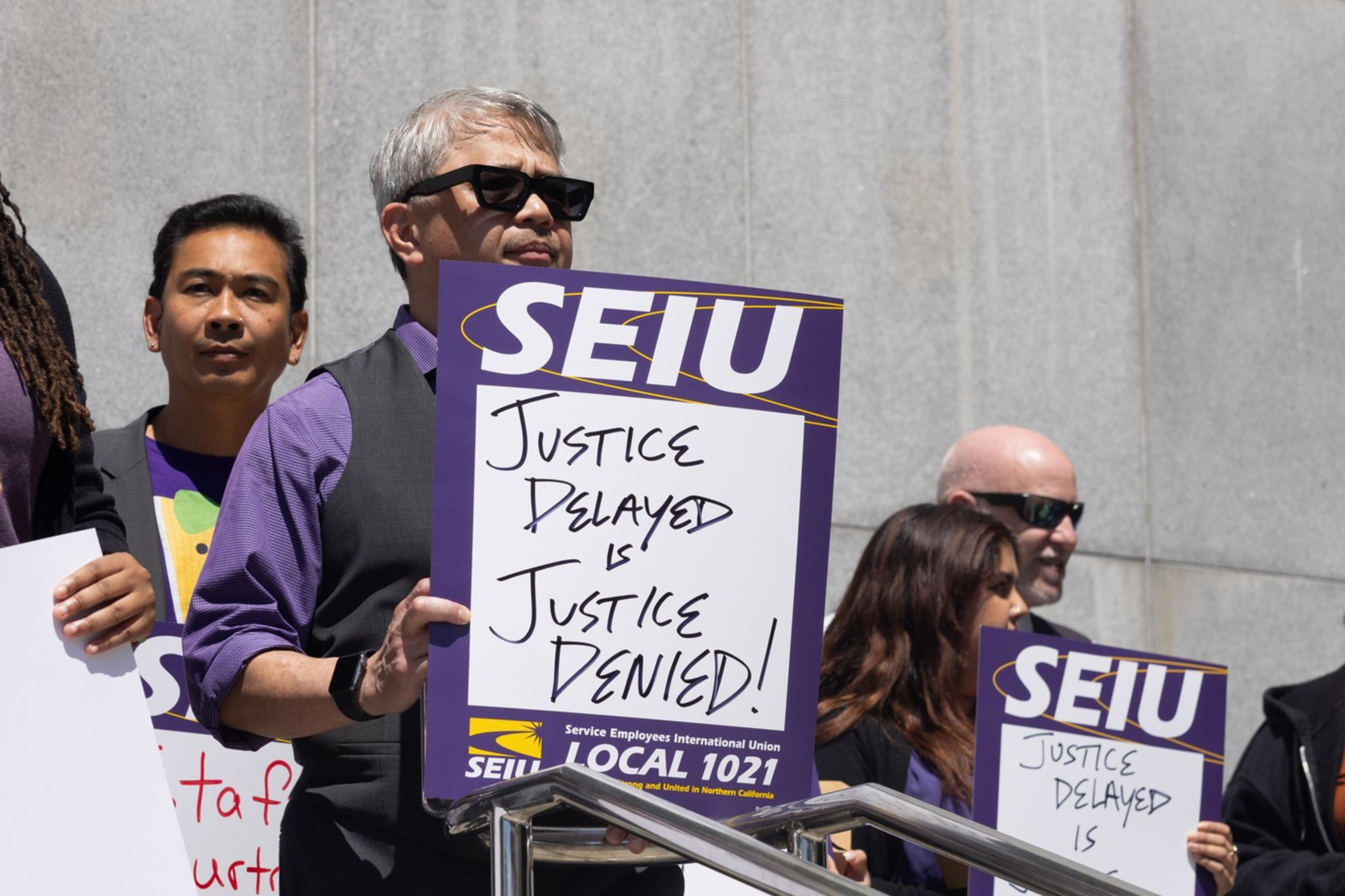 People stand on courthouse steps holding "Justice Delayed is Justice Denied!" signs under a plaque reading "Thomas J. Cahill Hall of Justice."