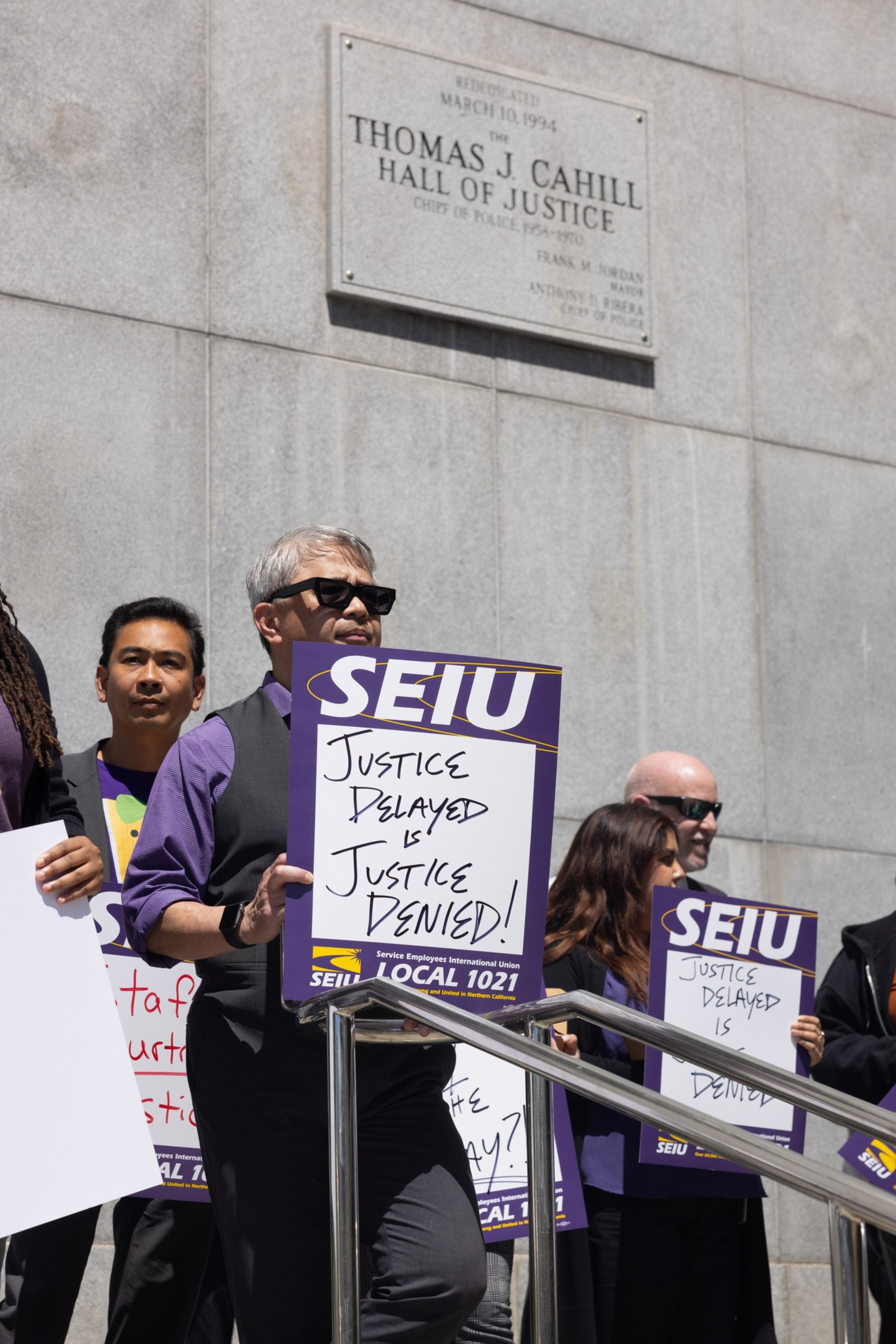People stand on courthouse steps holding "Justice Delayed is Justice Denied!" signs under a plaque reading "Thomas J. Cahill Hall of Justice."