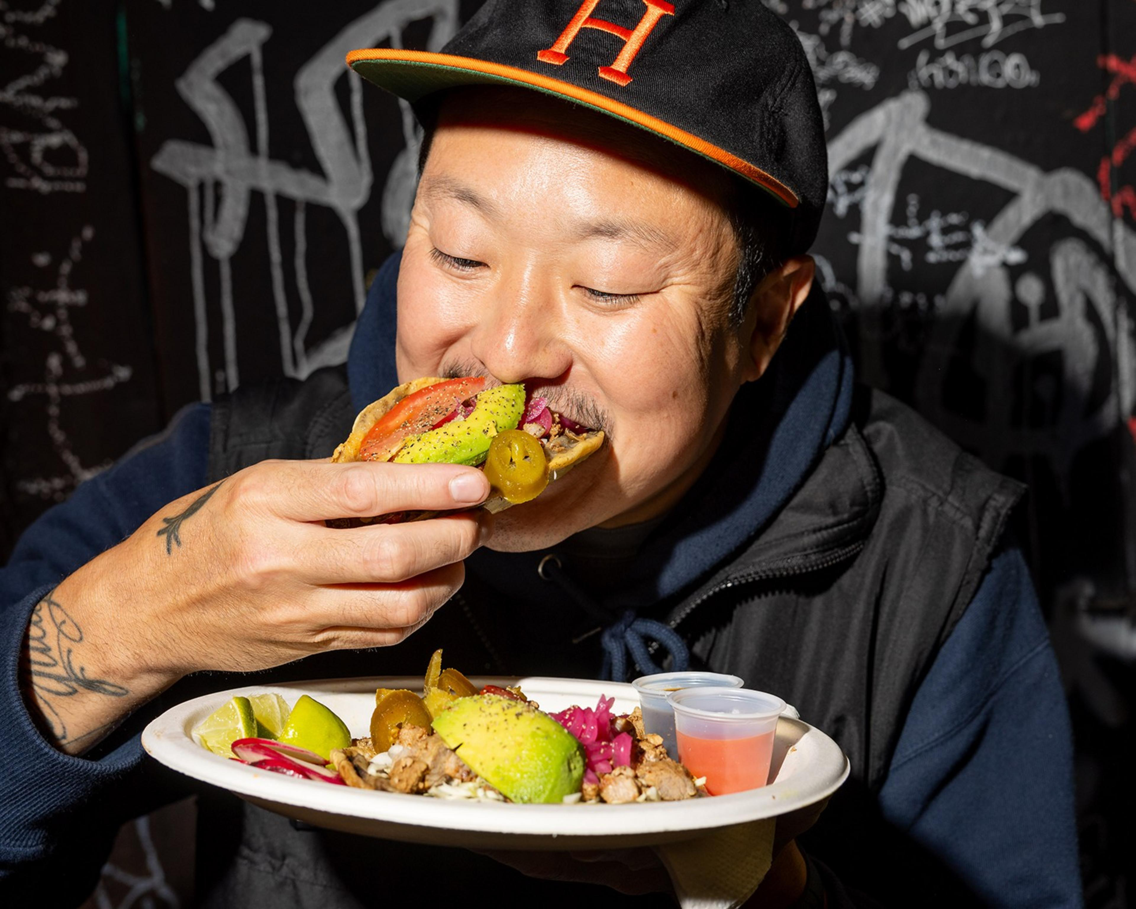 A man in a black hat is taking a bite of a taco while holding a plate with more food items, including avocado slices and a small container of sauce.