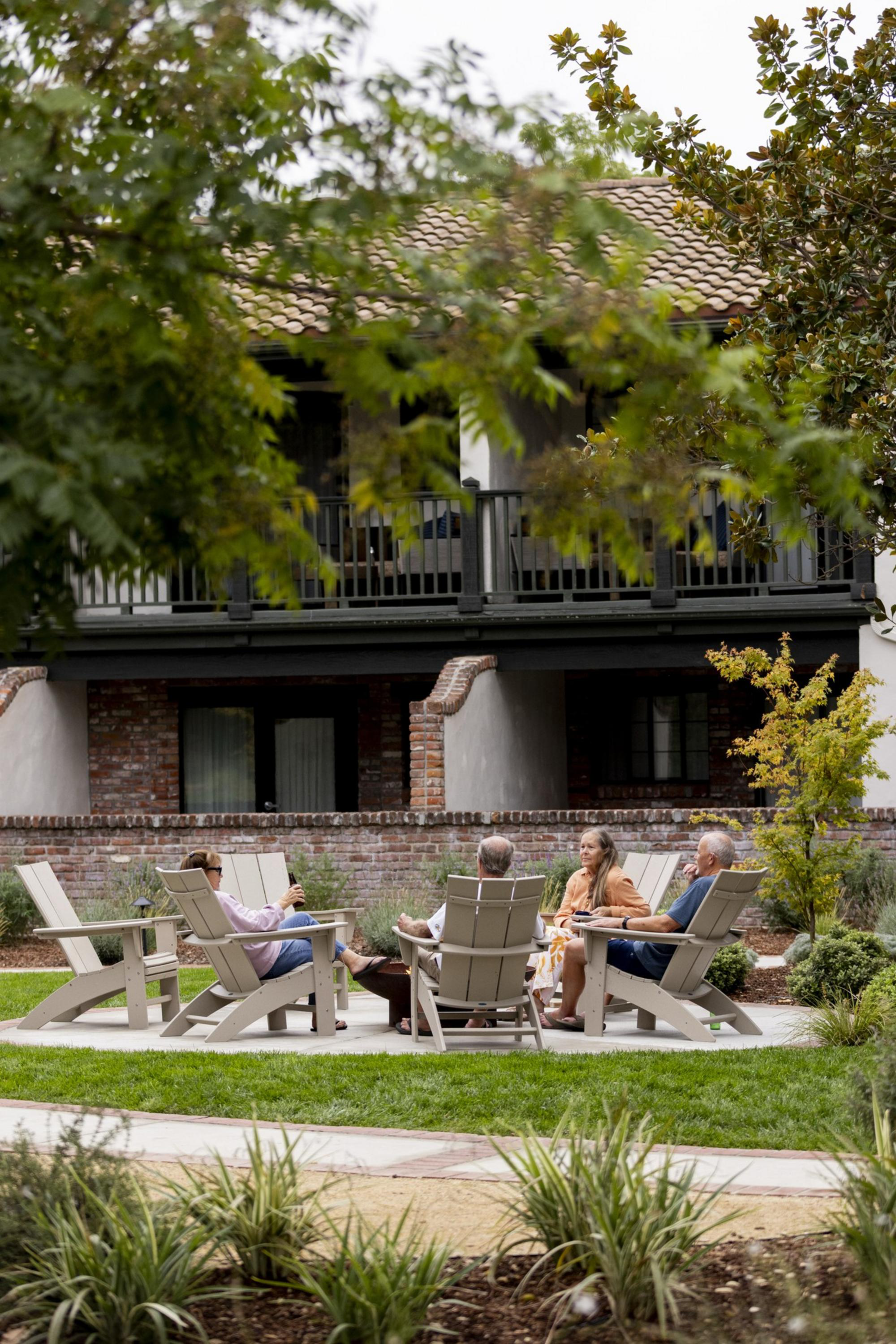 Four people sit in Adirondack chairs around a small table in a garden, with a brick building and balcony visible behind them.