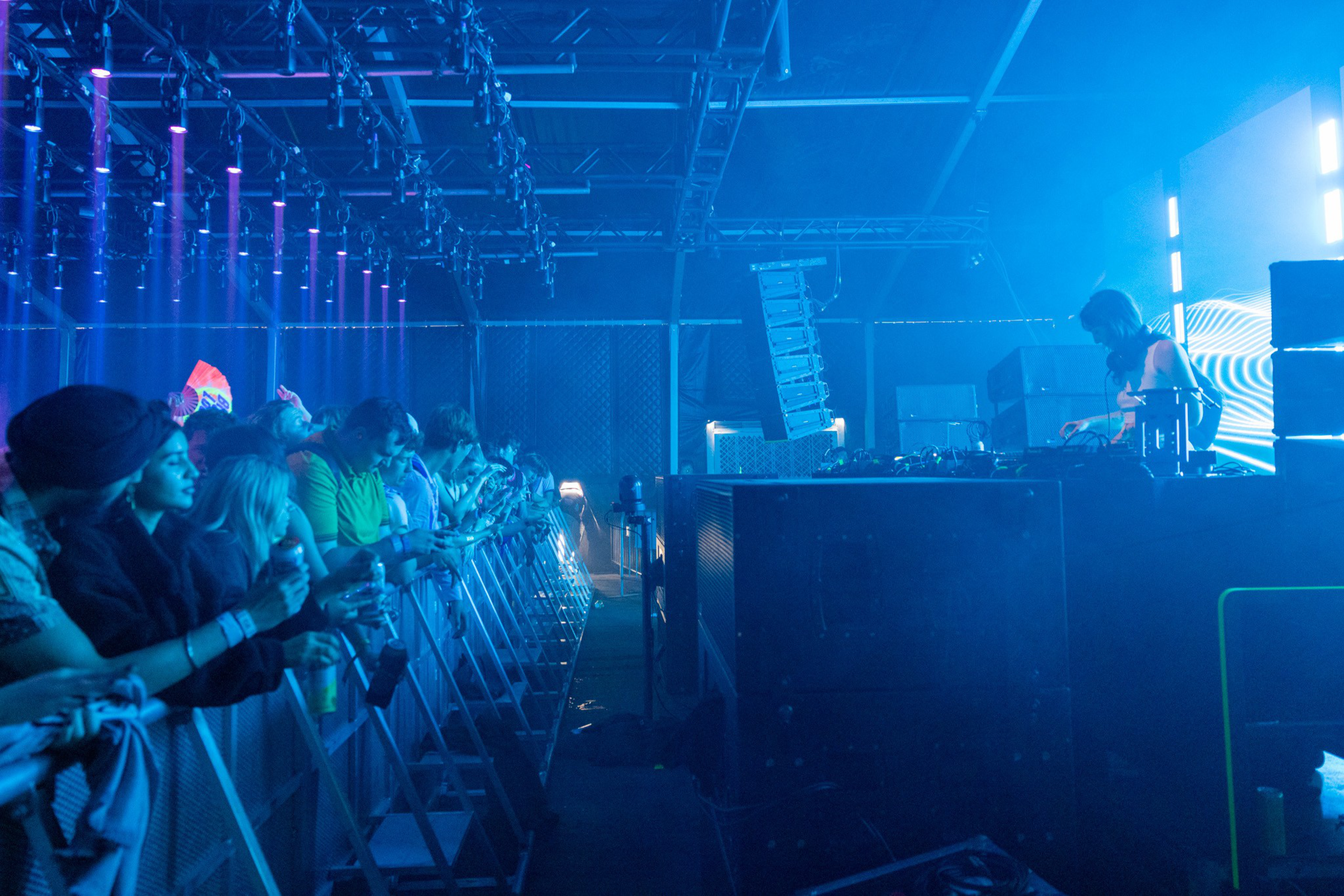 A DJ performs on an elevated stage with blue lighting while a crowd watches closely behind metal barricades in a dark indoor venue.