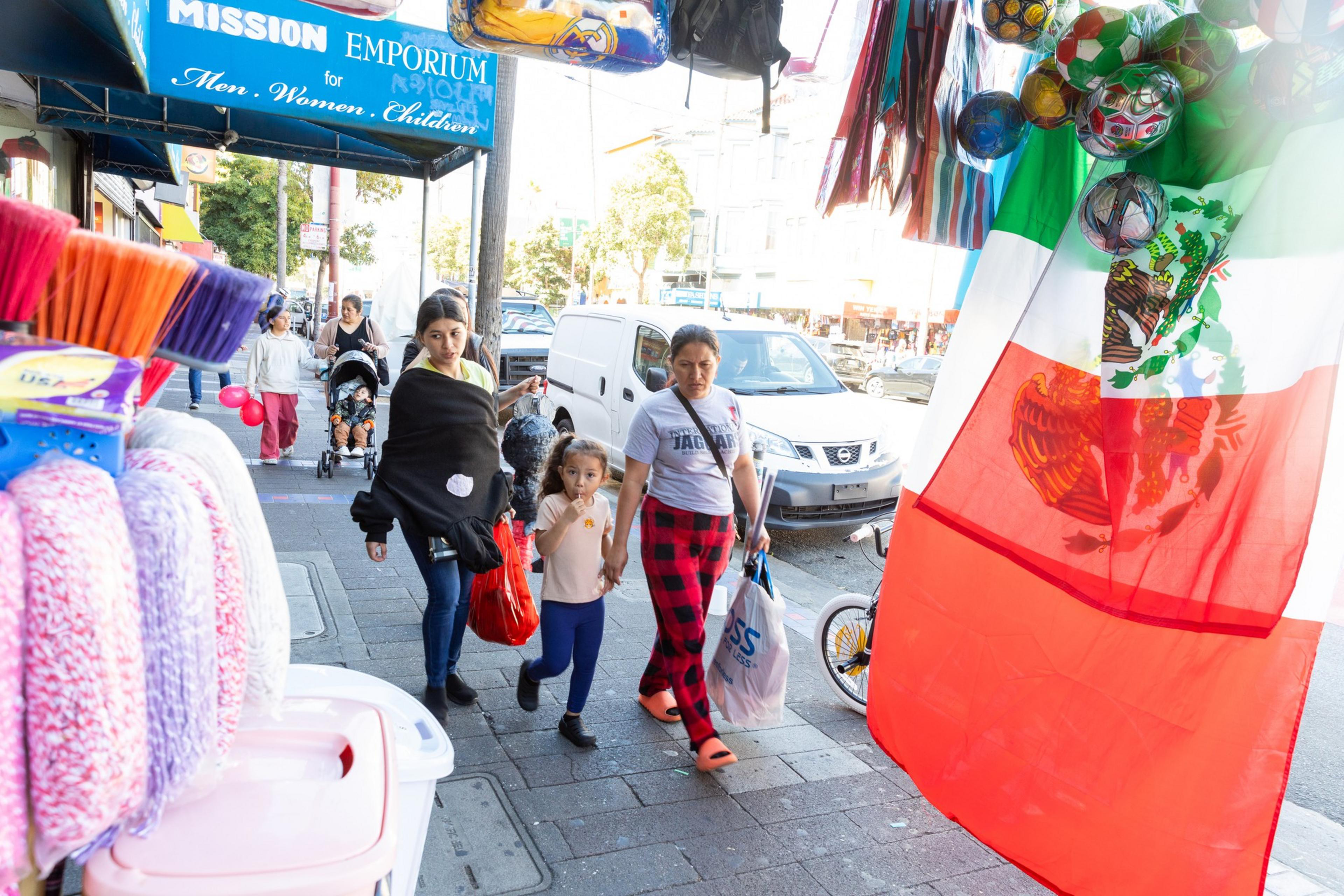A busy street scene shows people walking past colorful storefronts, a large Mexican flag, and a display of cleaning supplies. There are also balloons and soccer balls hanging above.