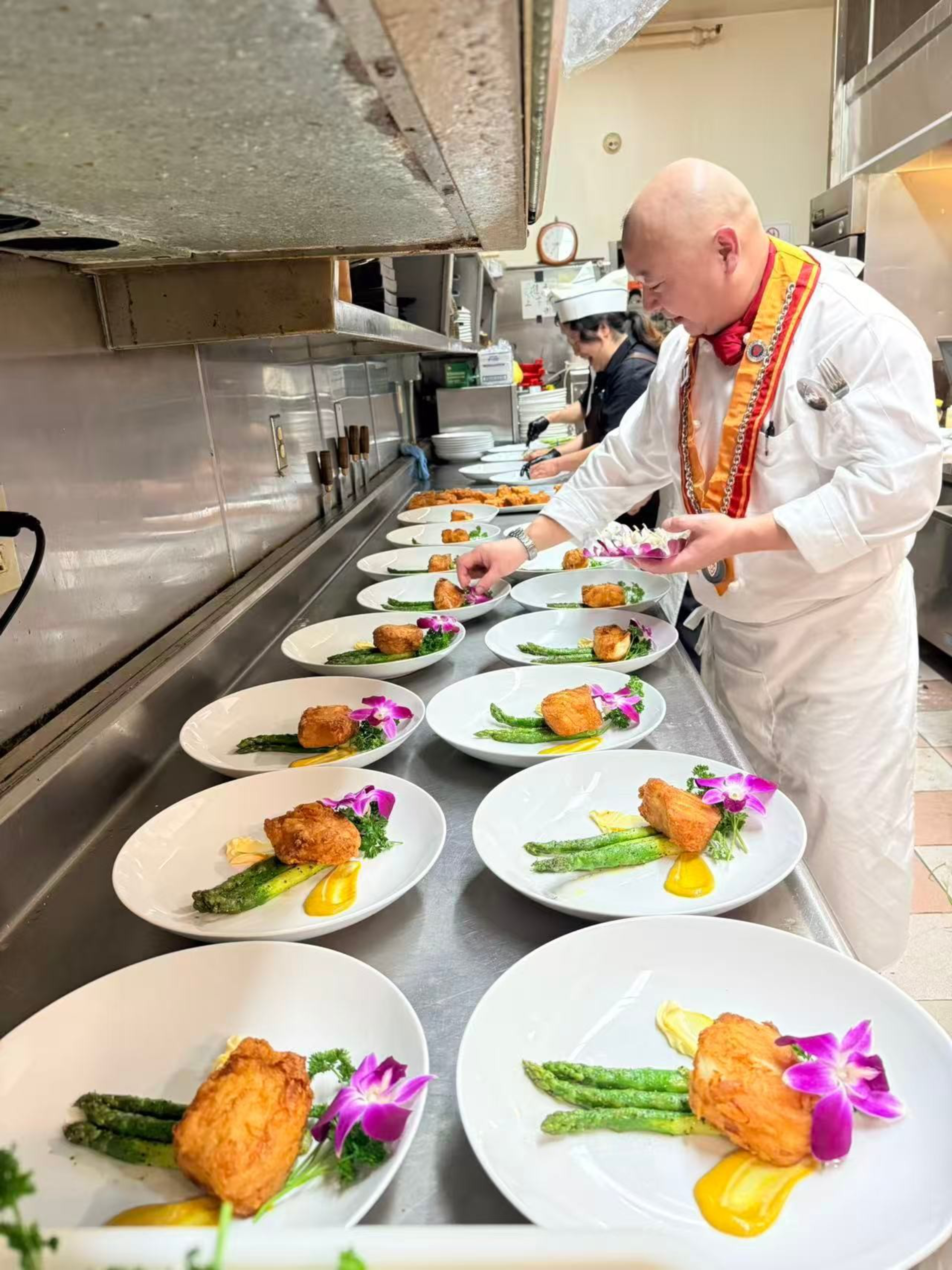 A chef is placing purple flowers on plates lined with cooked asparagus, a golden-brown fried item, and a yellow sauce in a restaurant kitchen.