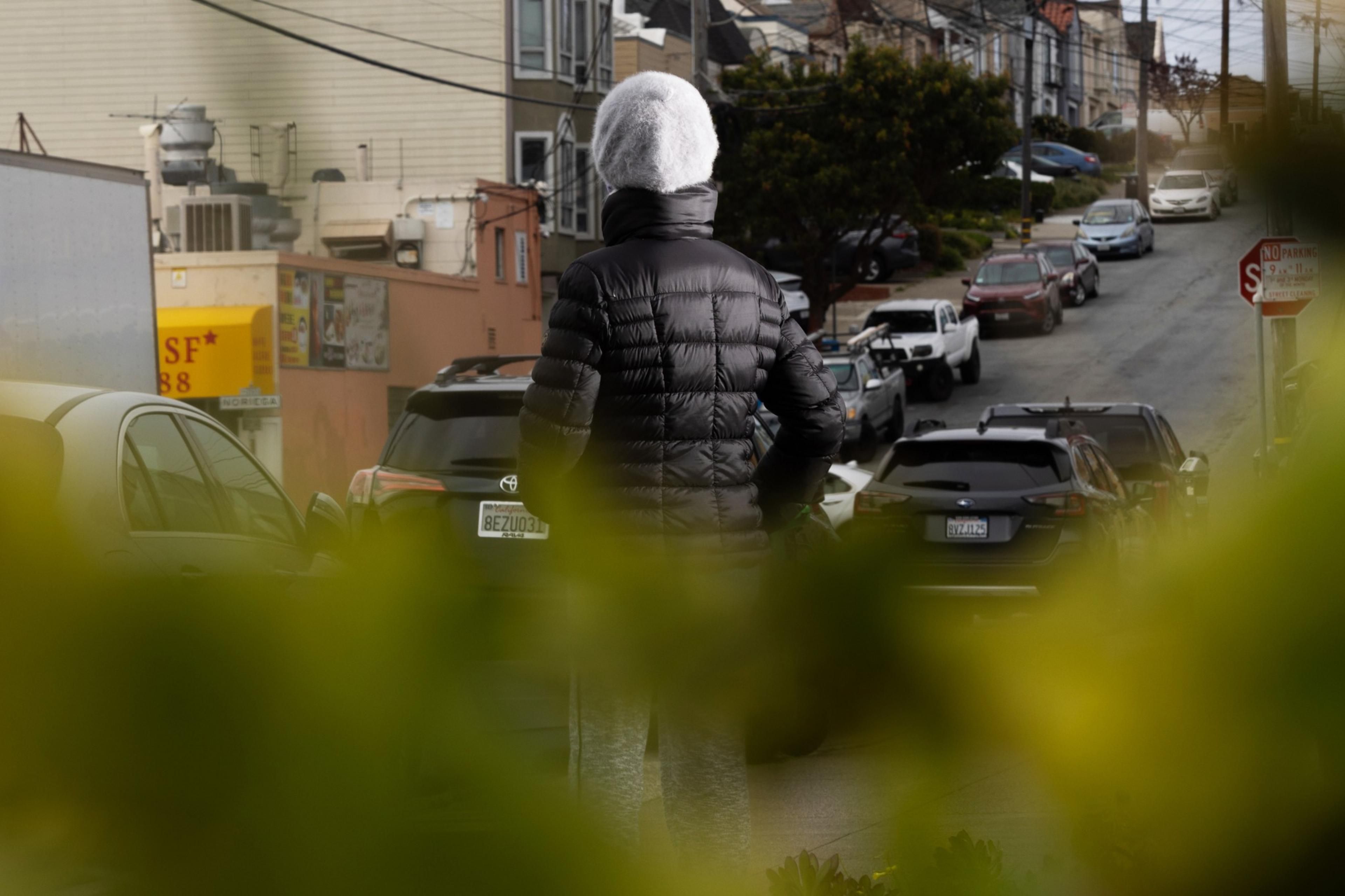A person in a black jacket stands on a street with parked cars, viewed through blurry foliage.
