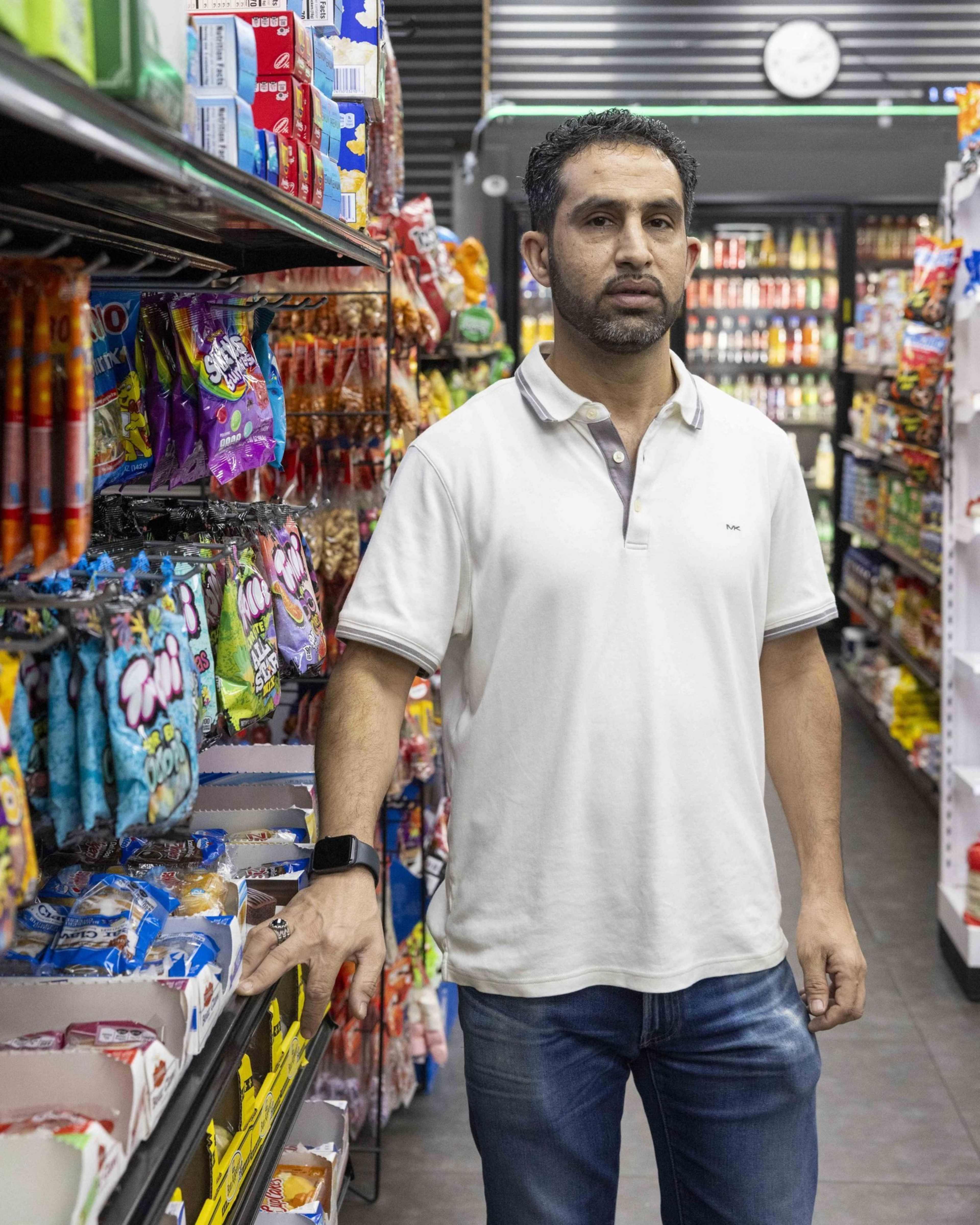 A man in a white polo shirt stands in a grocery aisle surrounded by colorful snack packages. He has a smartwatch on his wrist and a clock is visible behind him.