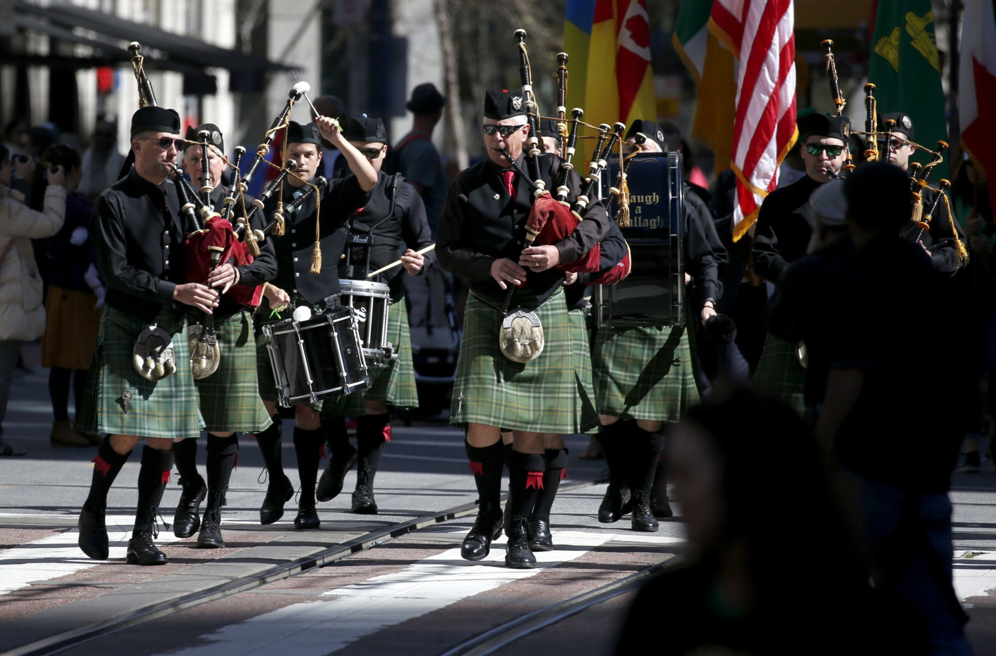 A marching band wearing green tartan kilts and black shirts plays bagpipes and drums while walking down a street with flags in the background.