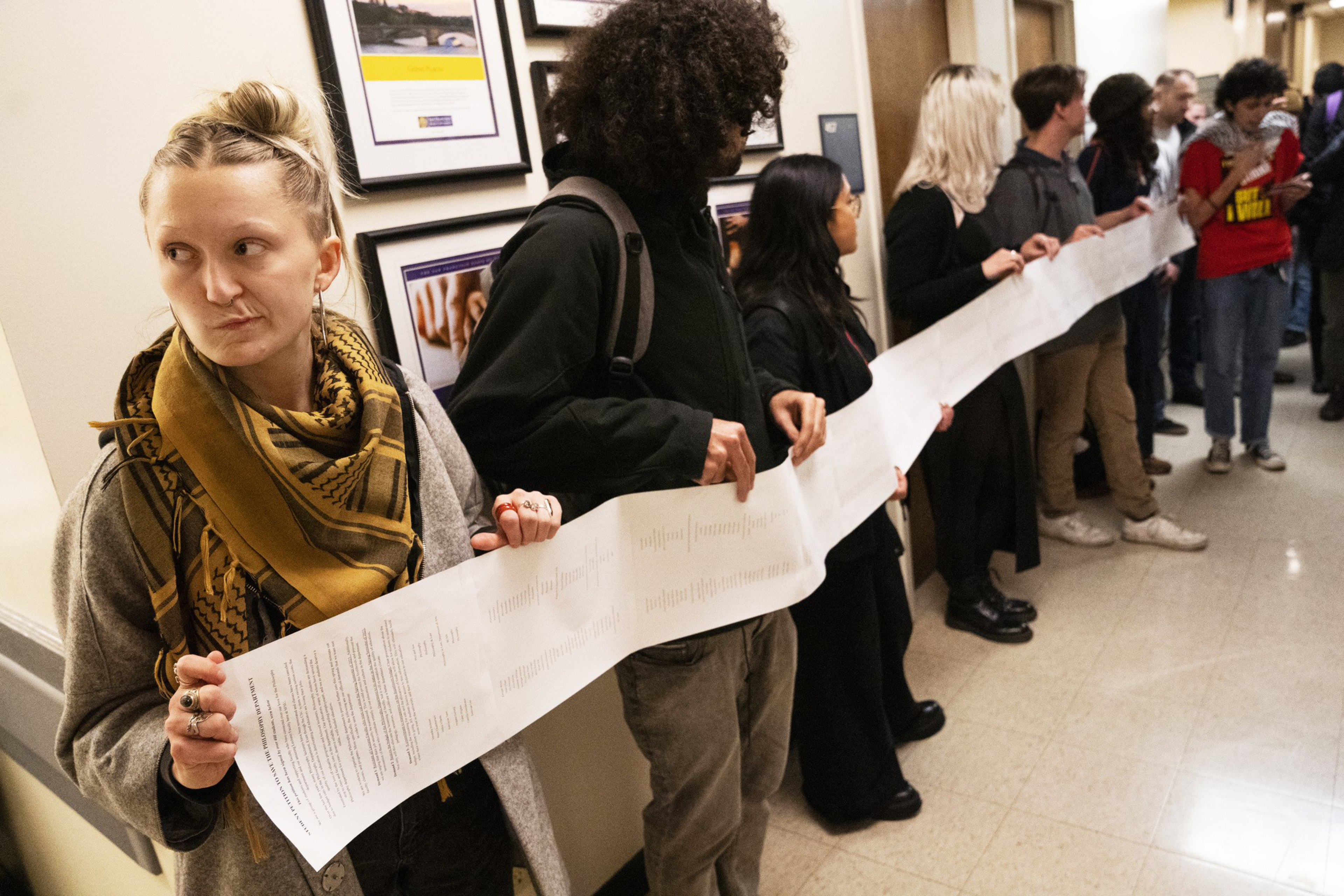 A group of people is standing in a hallway holding a long piece of paper. They appear to be part of a demonstration or protest, all facing one direction.