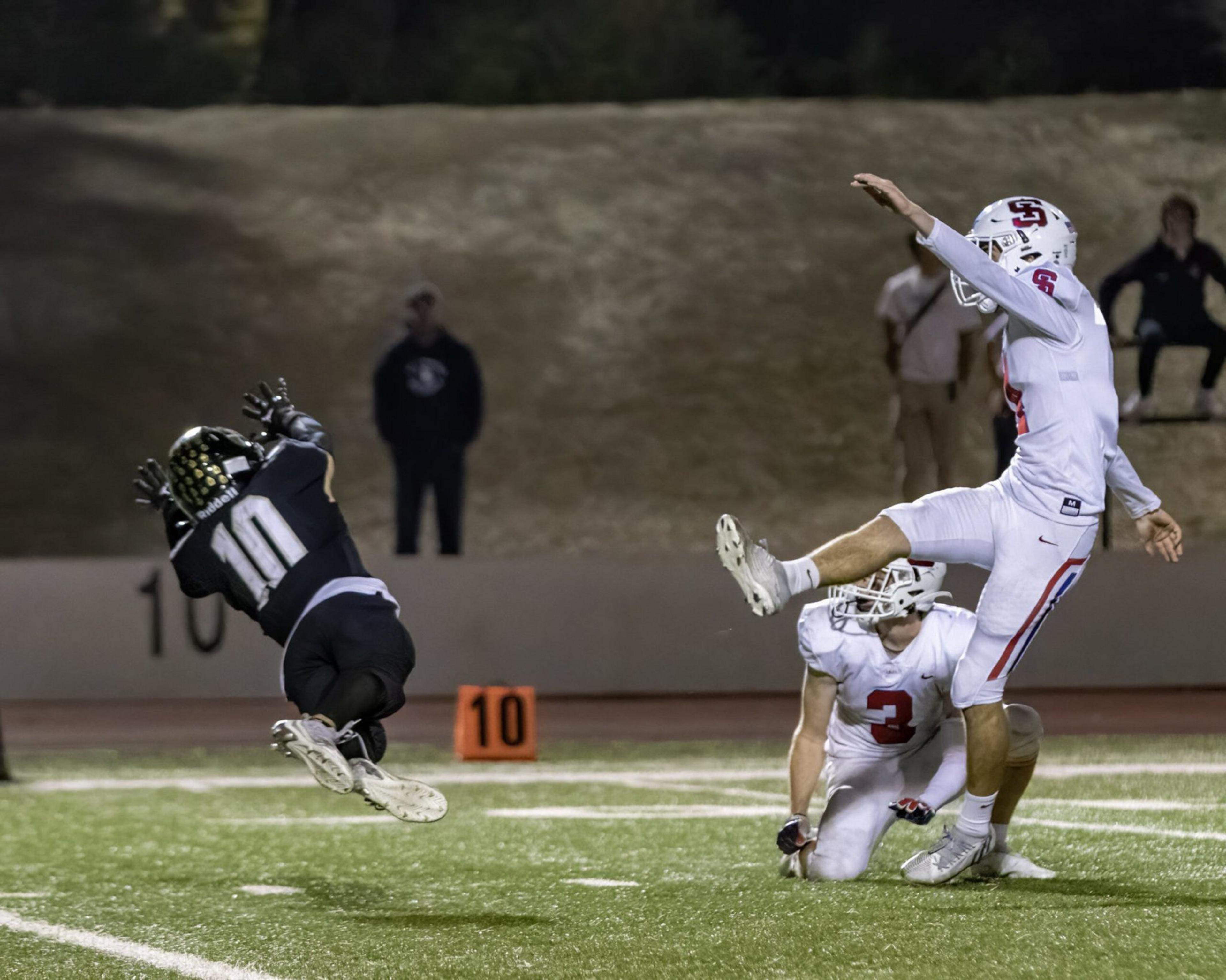 A football player in white kicks the ball as a teammate holds it, while a player in black leaps to block or catch it near the 10-yard line.