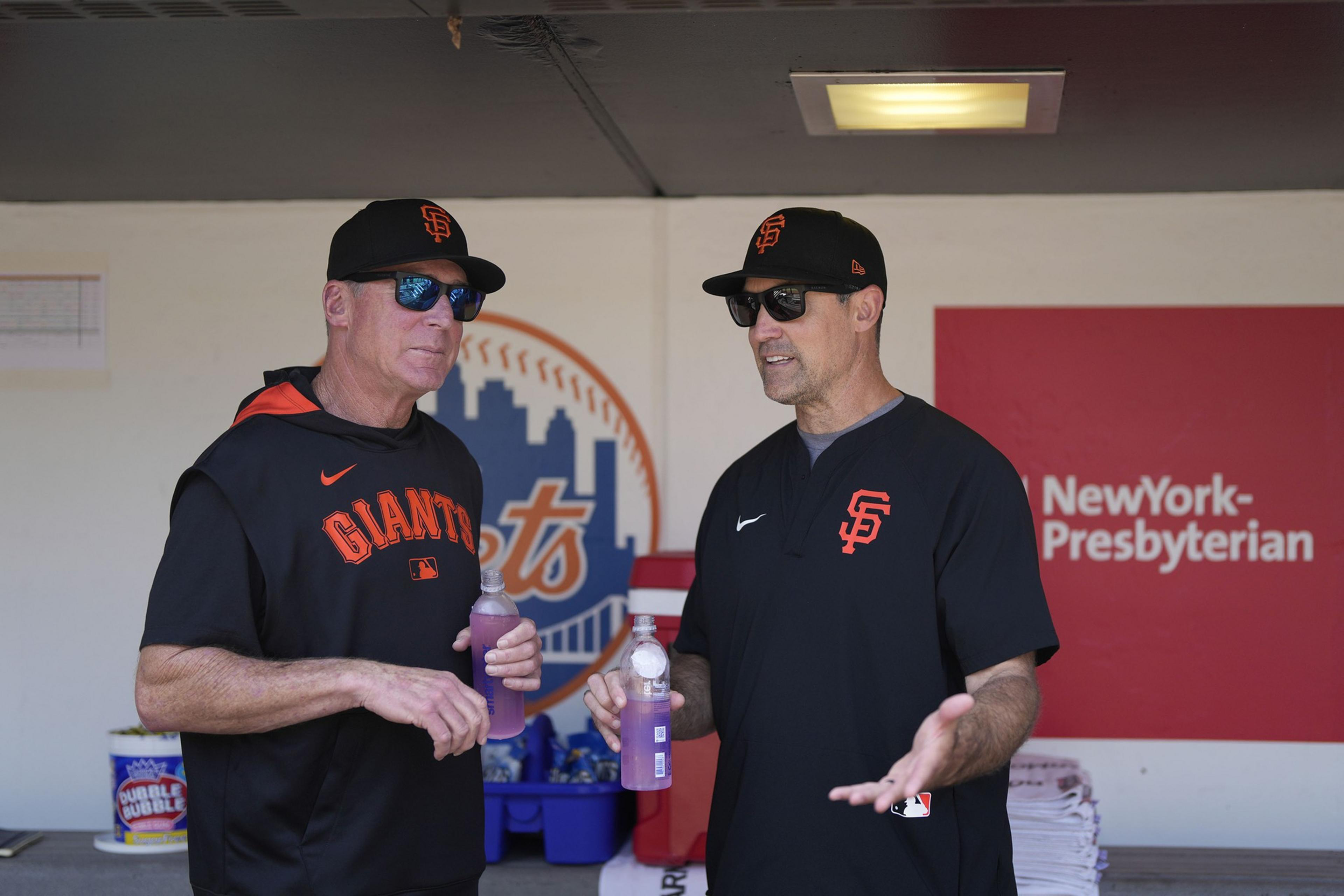 Two men in San Francisco Giants gear and sunglasses stand and chat in a baseball dugout, each holding a purple sports drink bottle.