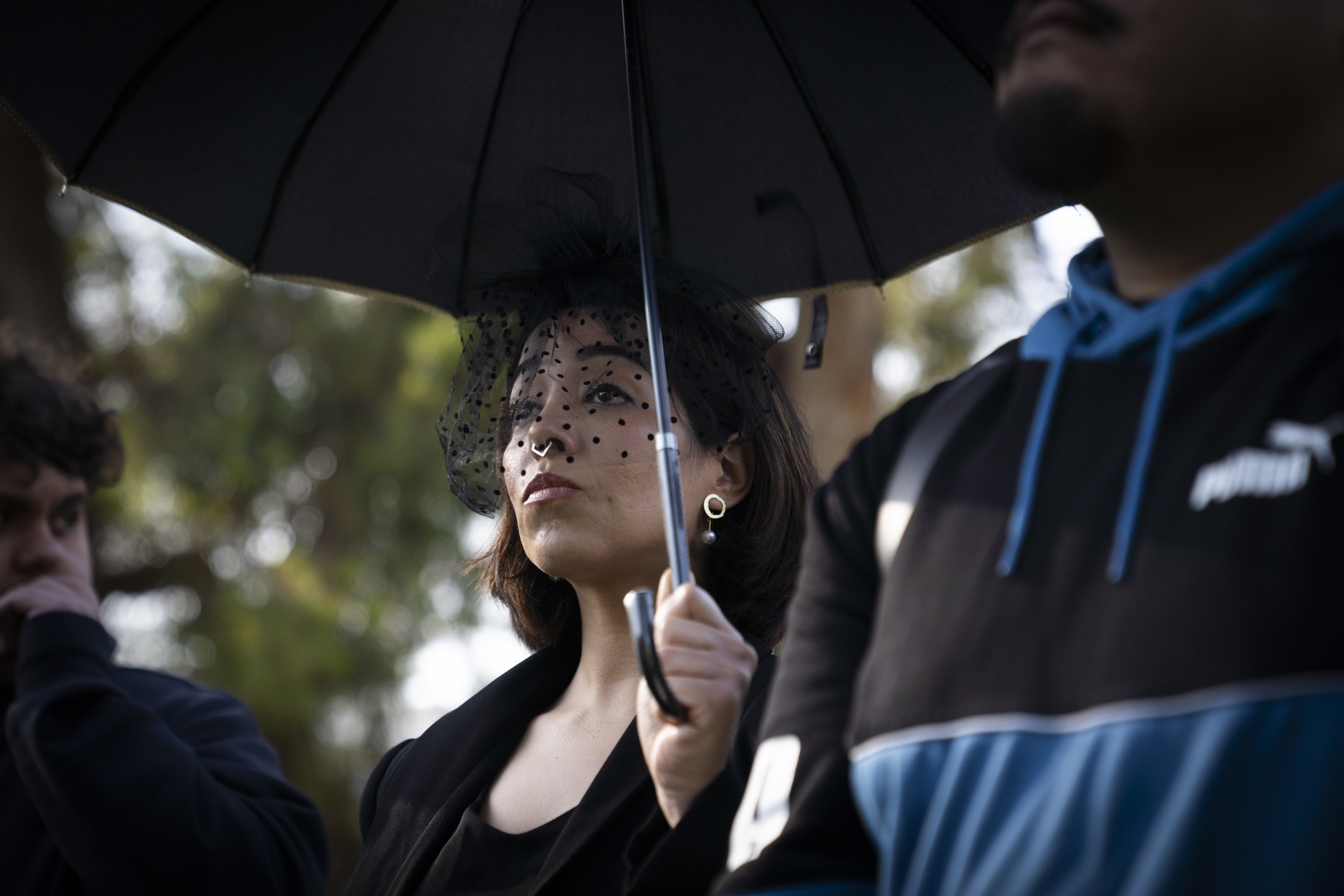 A woman holds a black umbrella, wearing a black veil with polka dots and pearl earrings. She stands among others, with blurred greenery in the background.