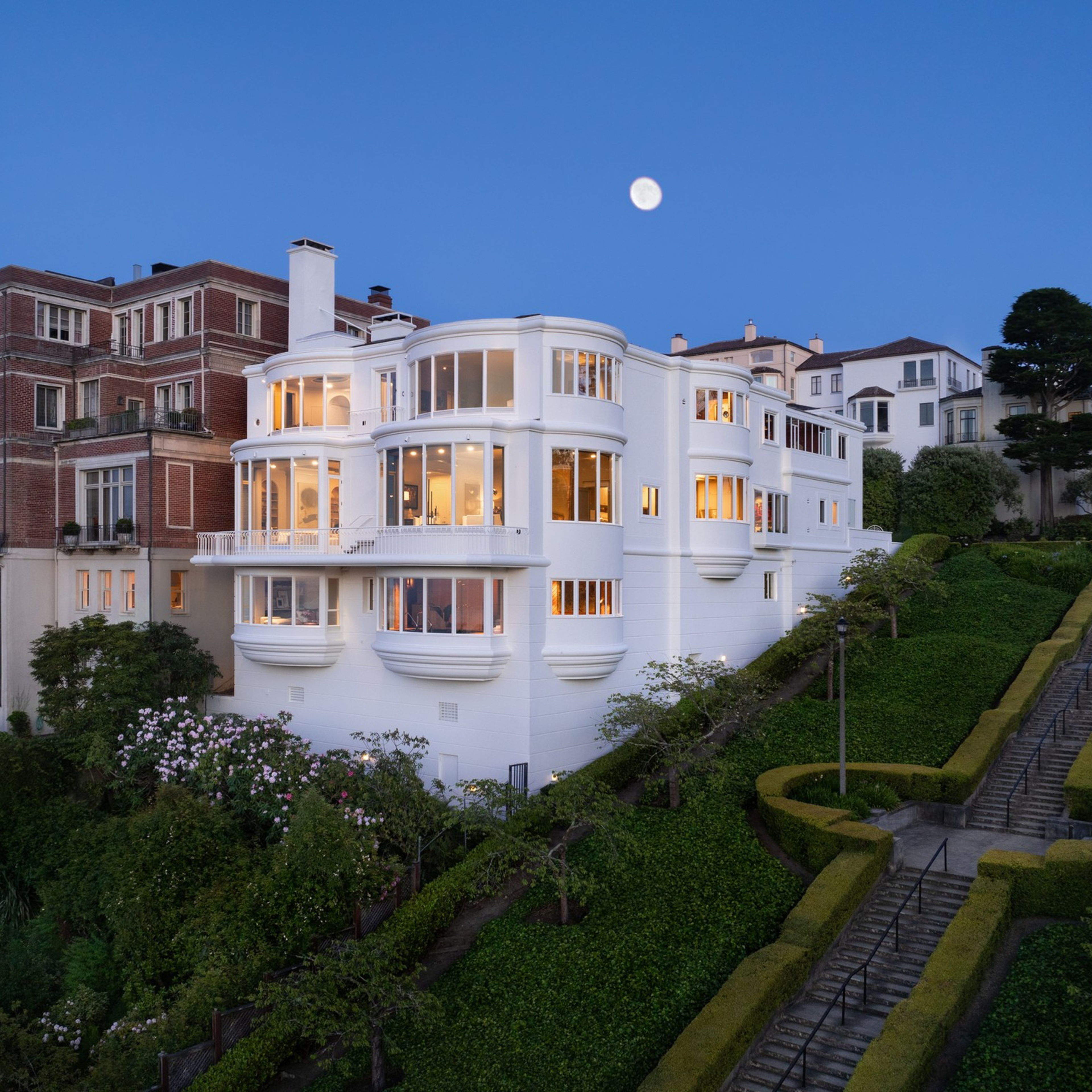 The image shows a large, elegant white house with multiple rounded, glass windows, lush greenery, and a staircase. It's situated in a scenic, upscale neighborhood at dusk.