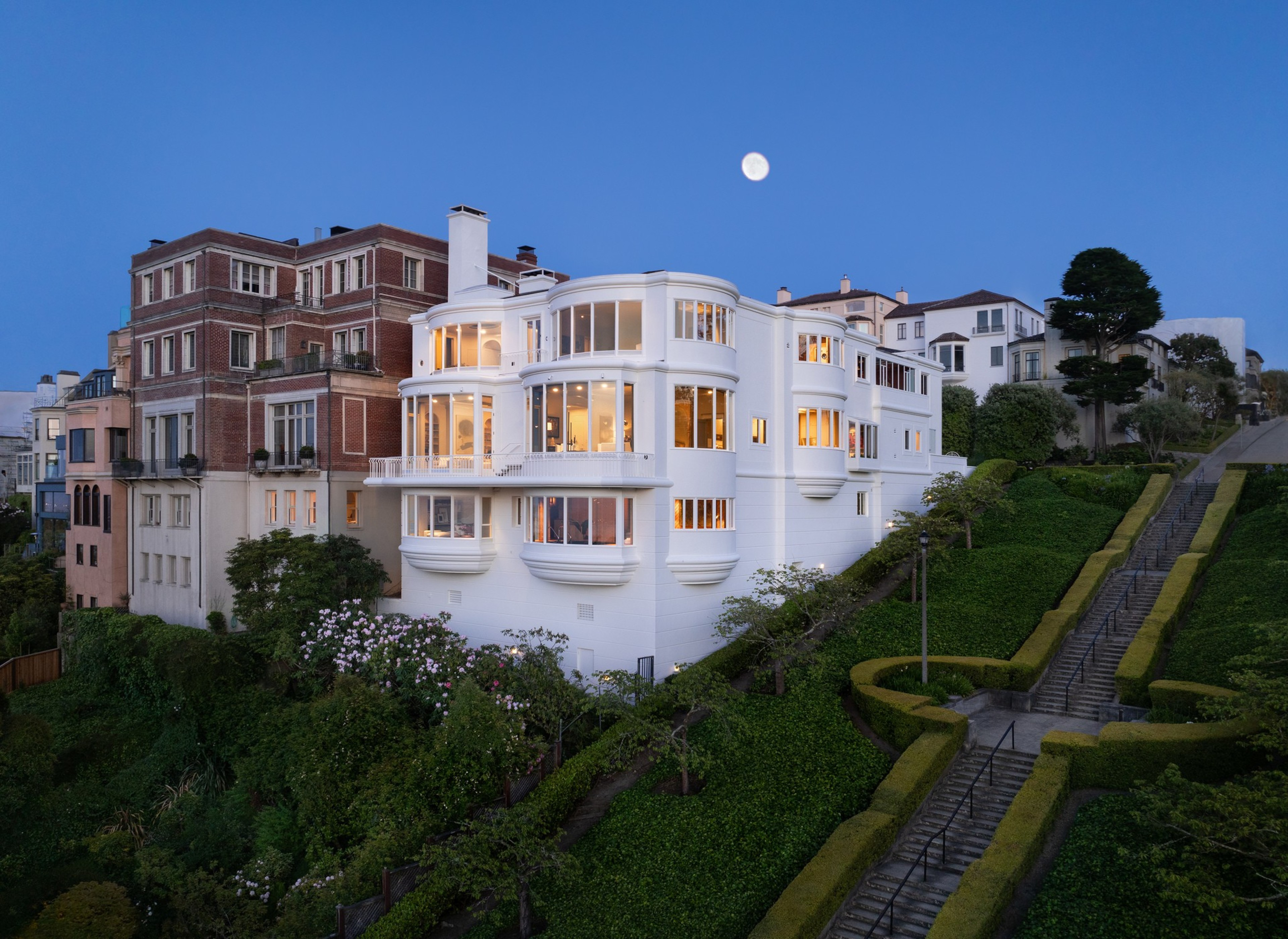 The image shows a large, elegant white house with multiple rounded, glass windows, lush greenery, and a staircase. It's situated in a scenic, upscale neighborhood at dusk.