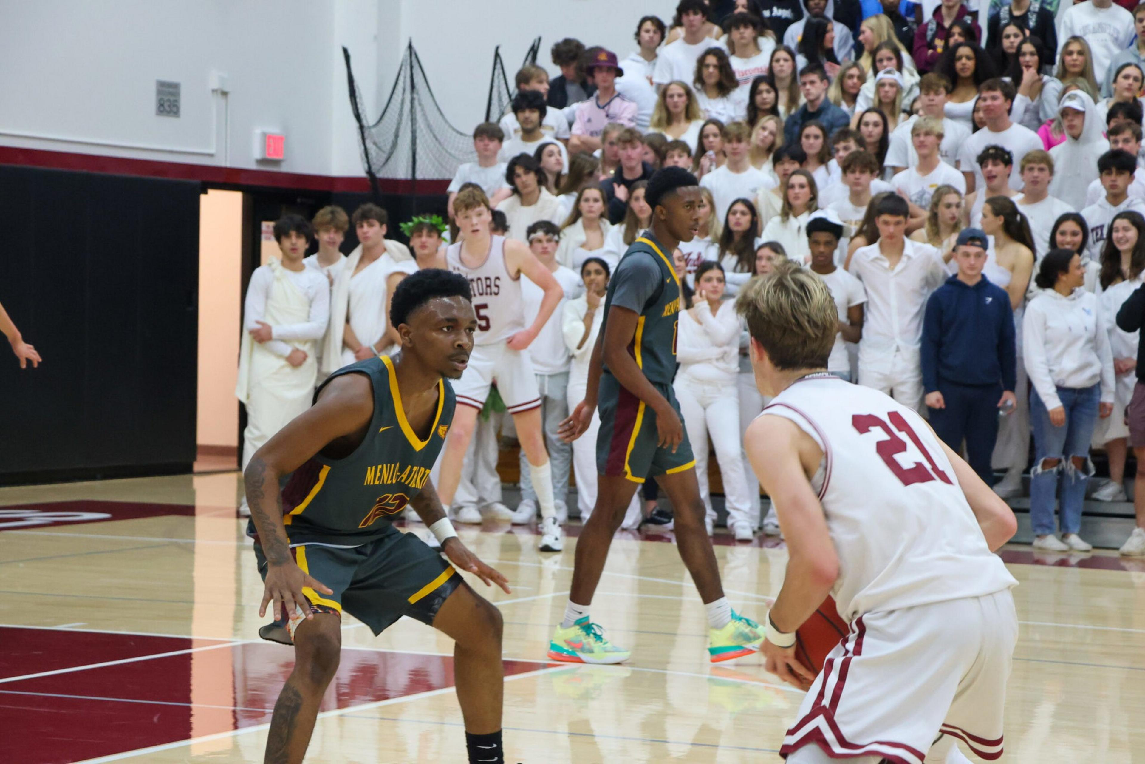 Two basketball players, one in white and one in dark jersey, face off on court while a large crowd dressed in white watches intently in the background.