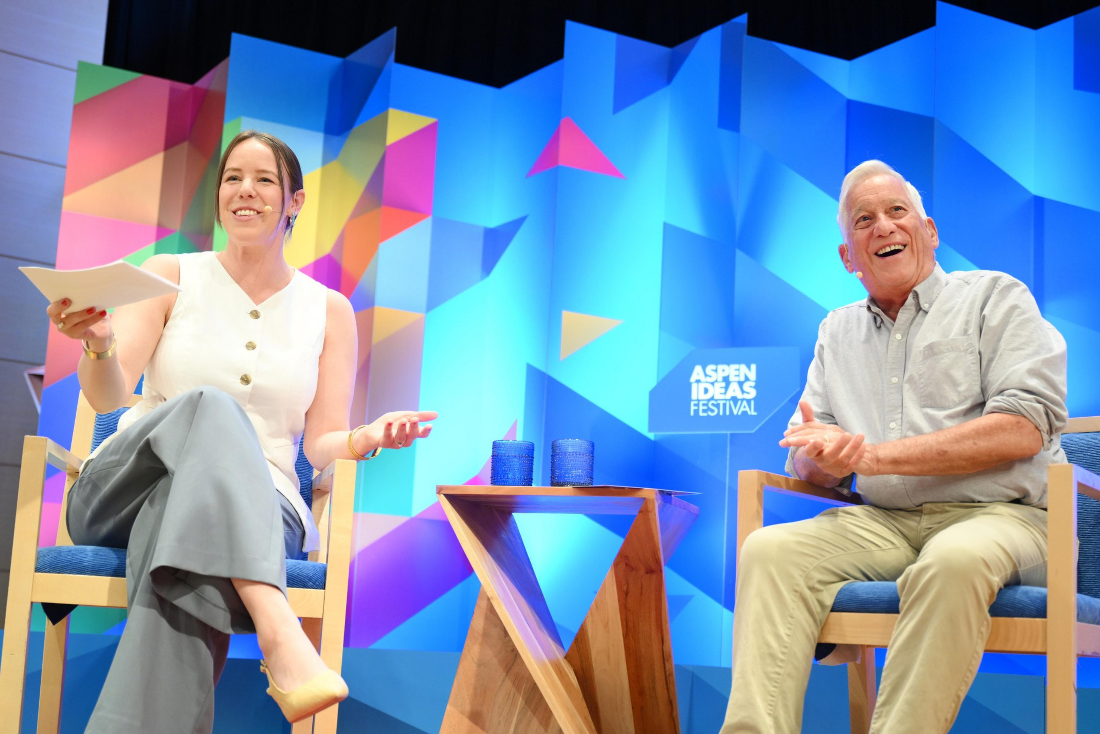 Host Sophie Bearman sits with journalist Walter Isaacson on stage in wooden chairs with a patterned blue background behind them.
