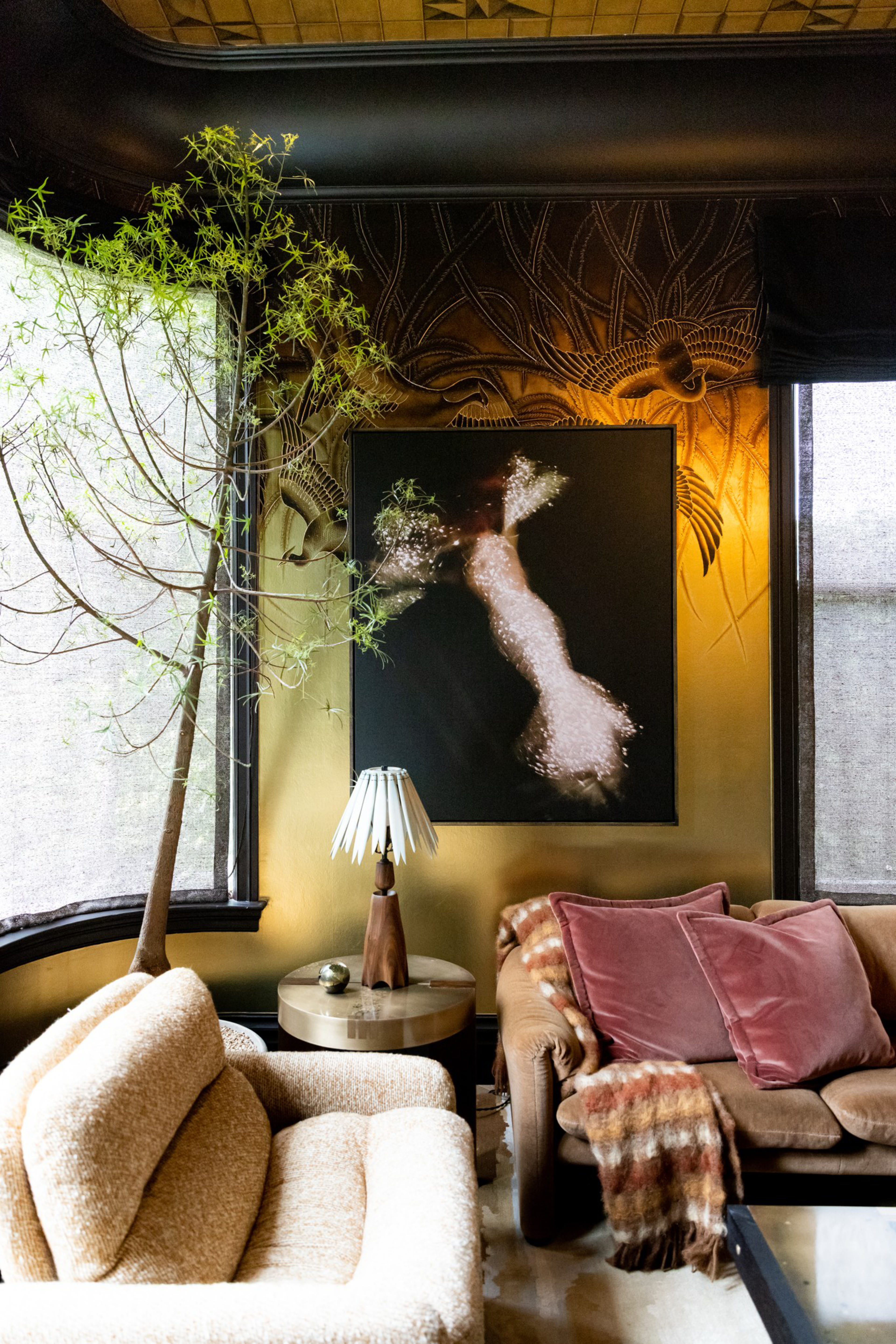 A cozy living room corner with a beige armchair, brown sofa with pink pillows and plaid throw, a tall potted plant, round side table with lamp, and dark abstract art.