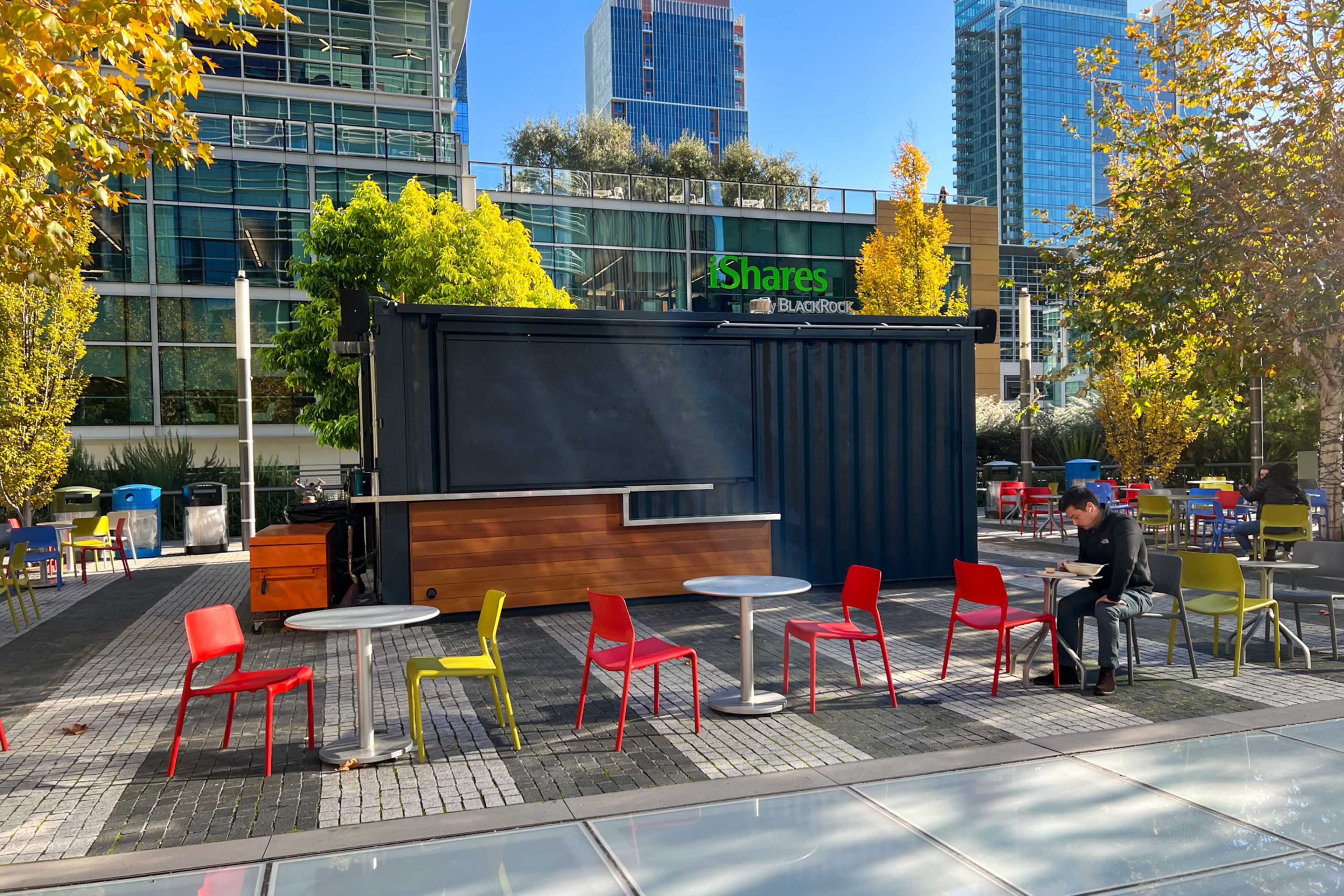 An outdoor seating area with colorful chairs and round tables sits in front of a closed black kiosk, surrounded by trees and tall buildings in daylight.