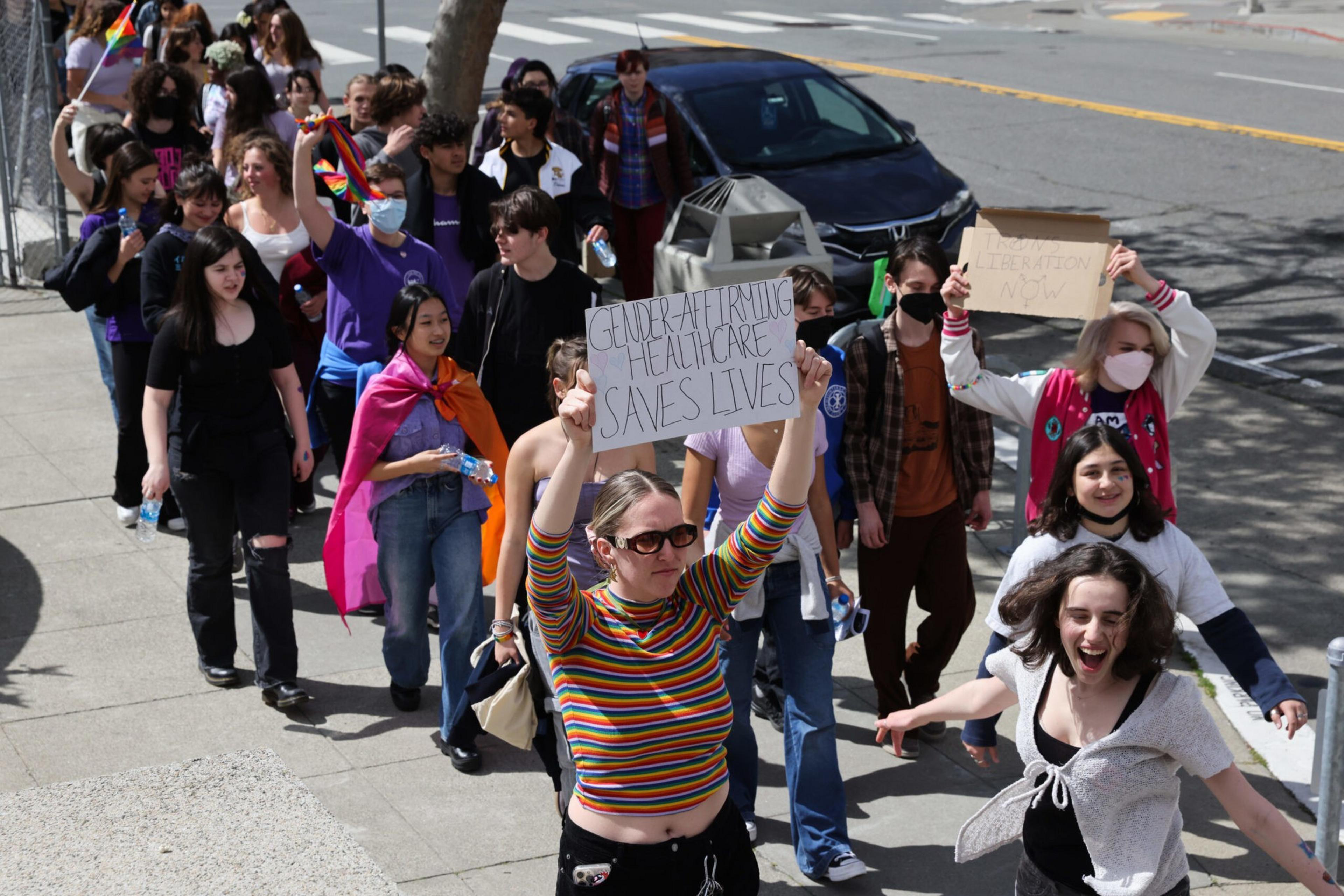 A diverse group of people march down a sidewalk, some holding LGBTQ+ flags and signs advocating for gender-affirming healthcare and abortion rights.