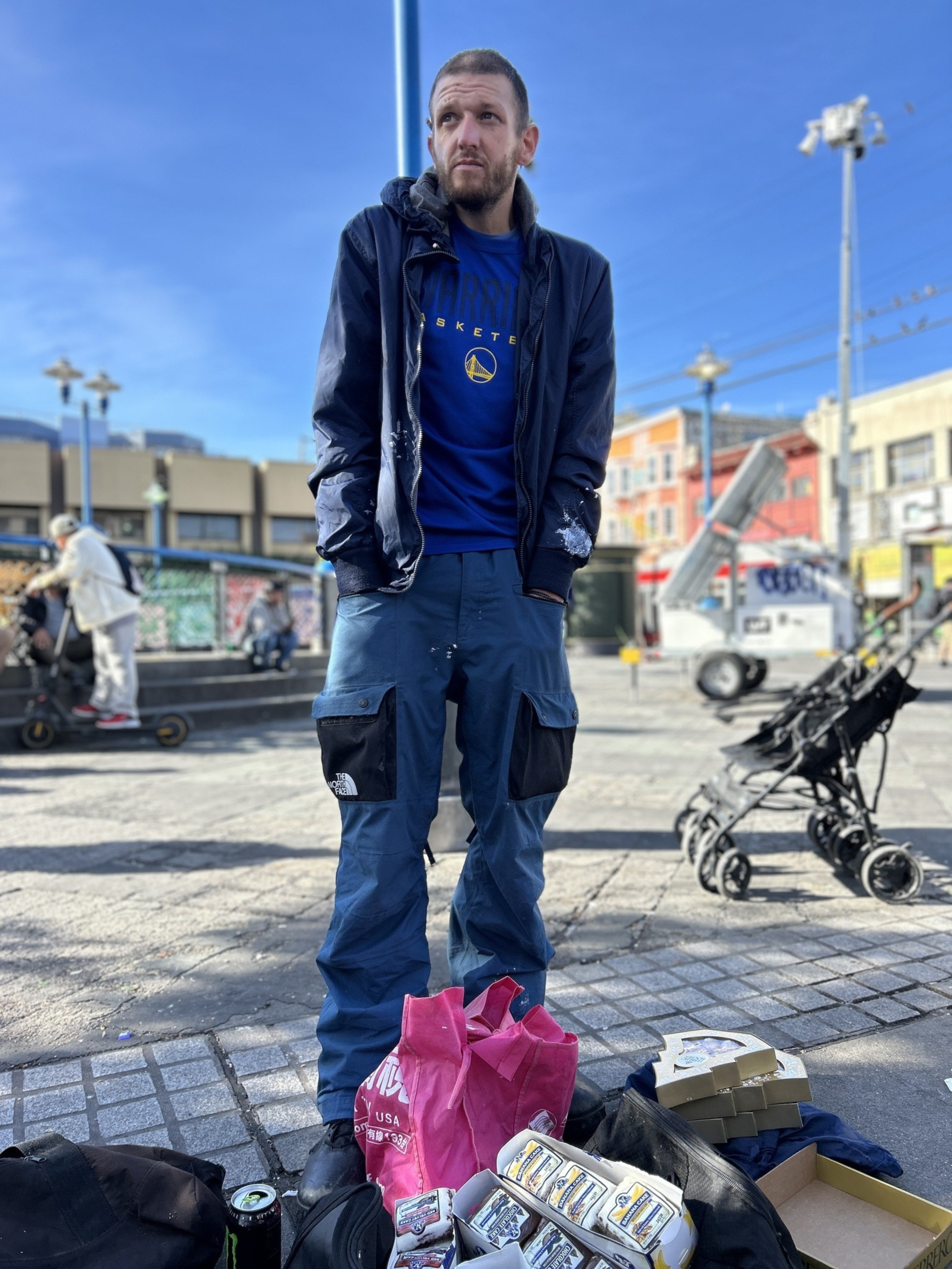 A man in a blue jacket and pants stands on a busy street, surrounded by bags and boxes. Behind him, there's a stroller and colorful buildings.