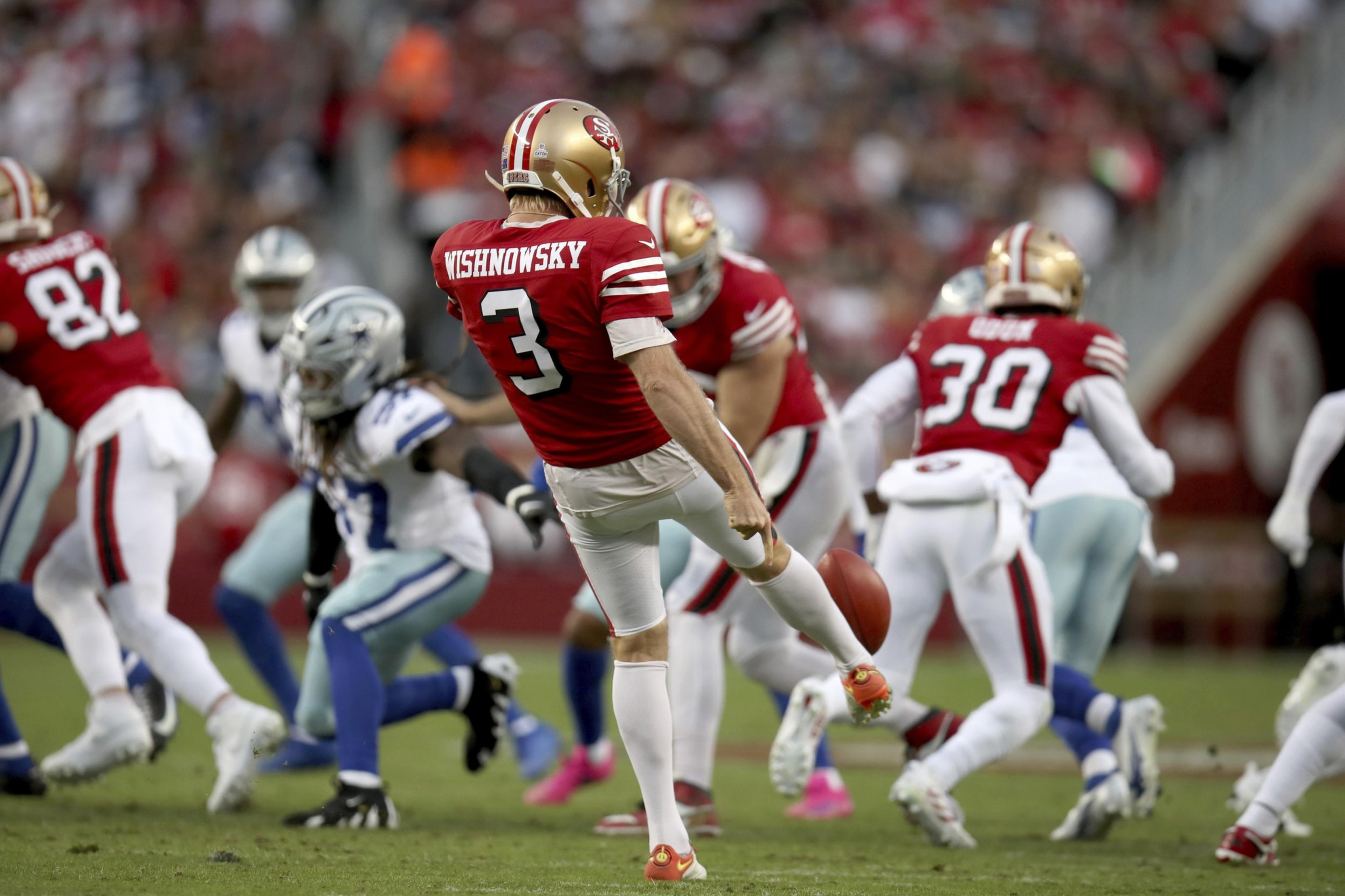 A football player in a red and gold uniform is kicking the ball, surrounded by teammates and opponents in blue and white. The crowd in the background is blurry.
