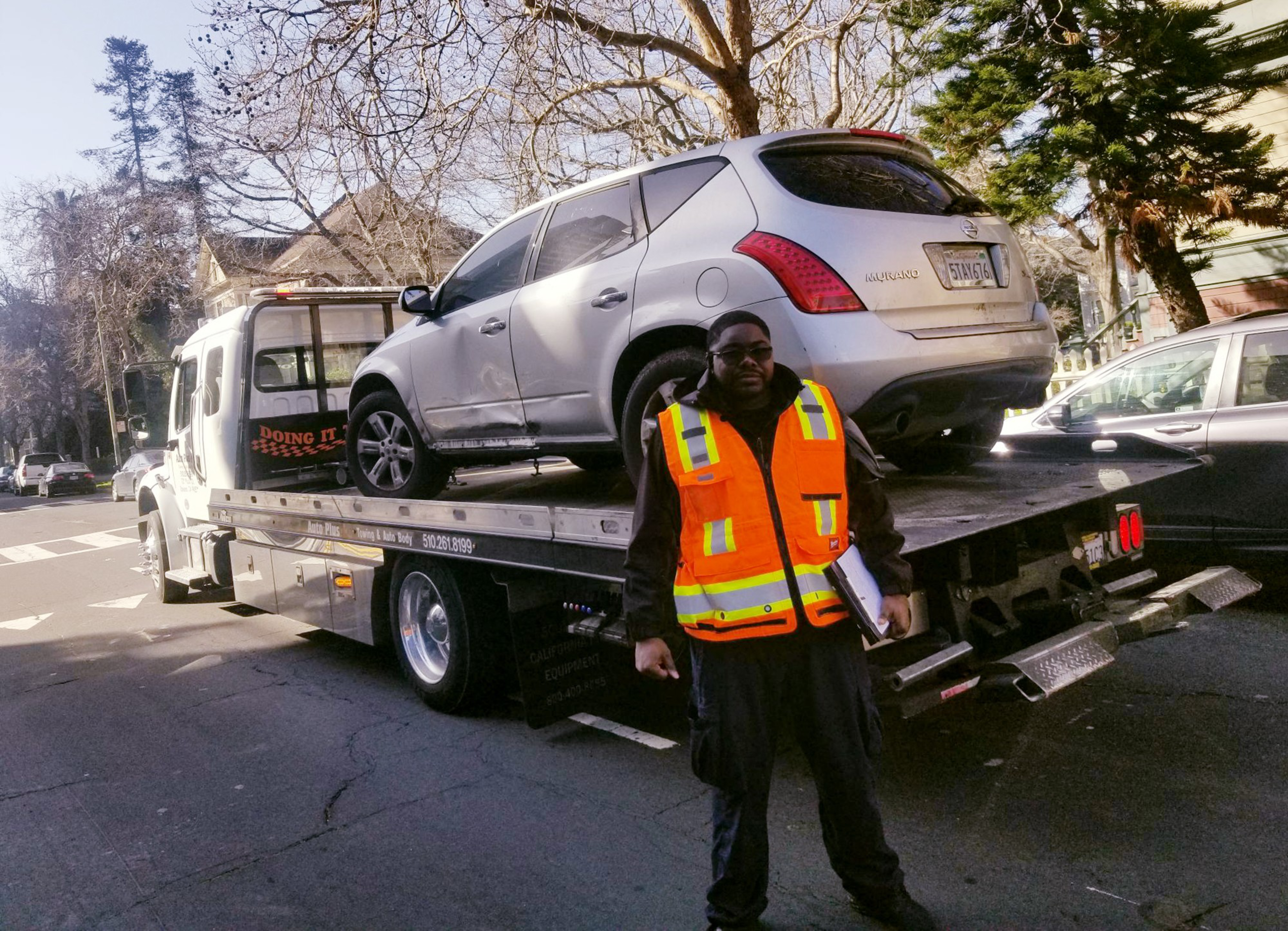A man in a reflective orange safety vest stands in front of a silver SUV loaded on a flatbed tow truck on a city street.