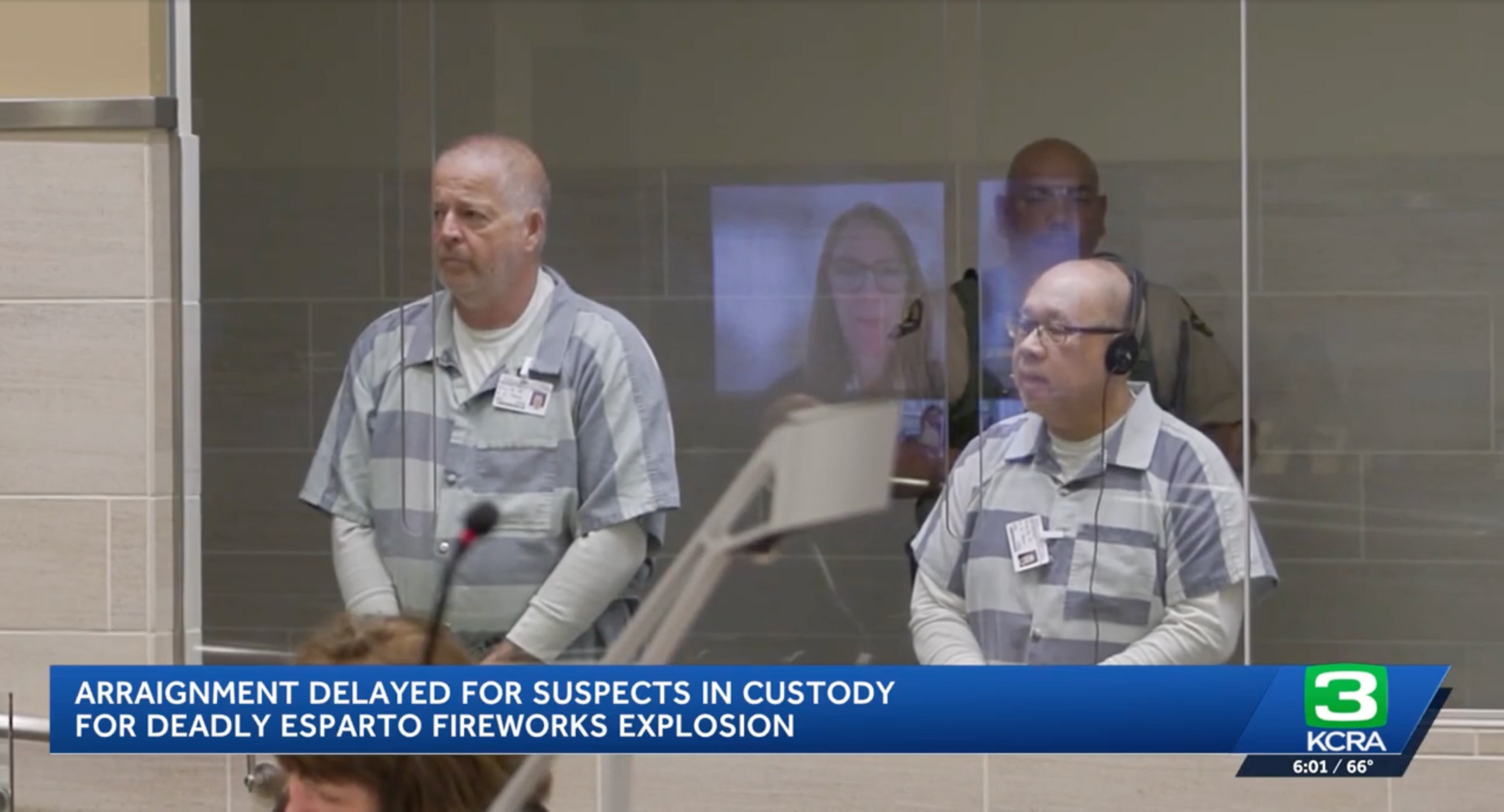 Two men in striped prison uniforms stand behind glass in a courtroom, with a guard visible behind them and a video call screen reflecting nearby.